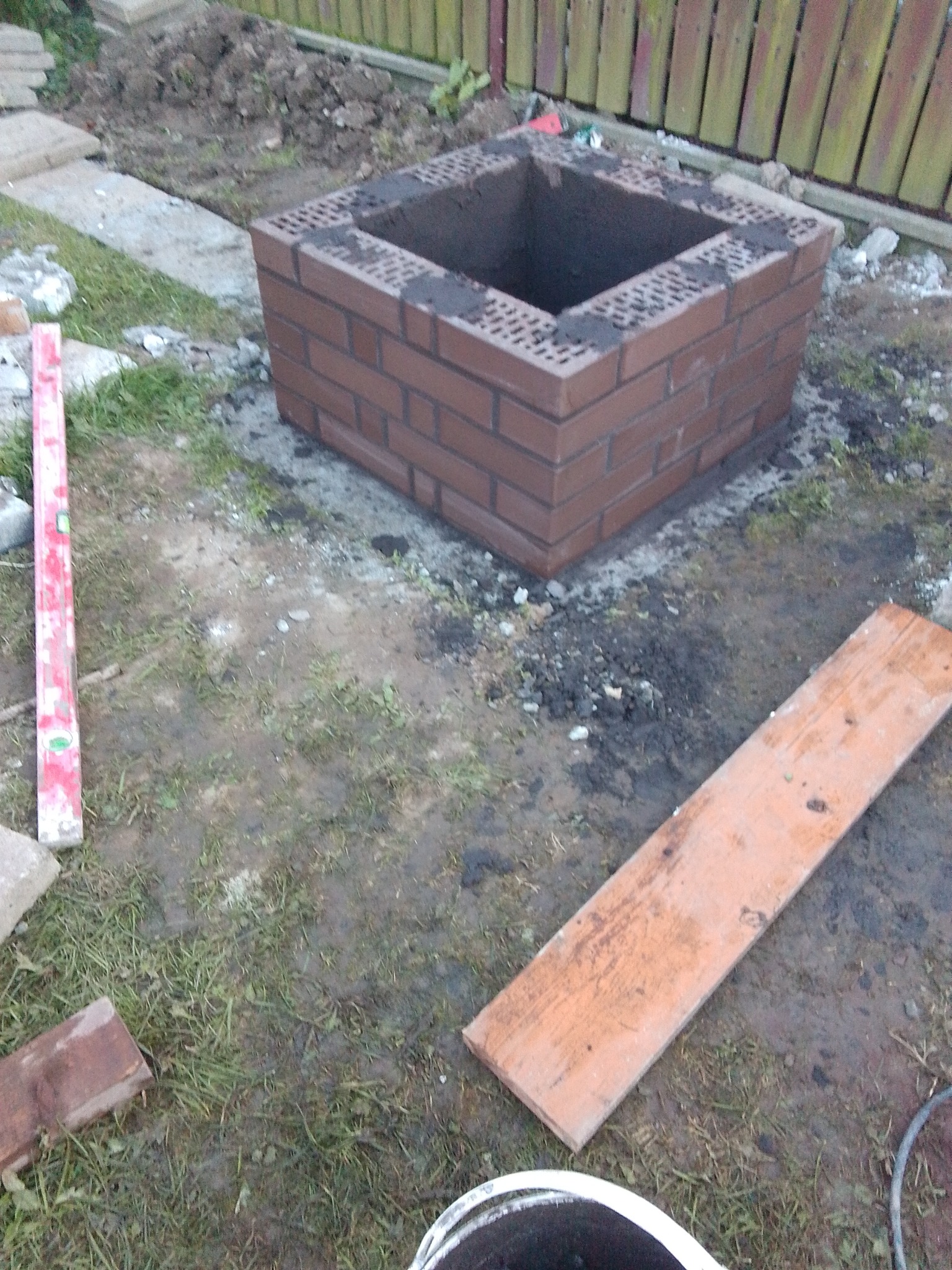 Murowana, kwadratowa konstrukcja z cegły klinkierowej w ogrodzie, prawdopodobnie pod palenisko lub grill, widoczna poziomica i deska, otoczenie w trakcie prac ziemnych.