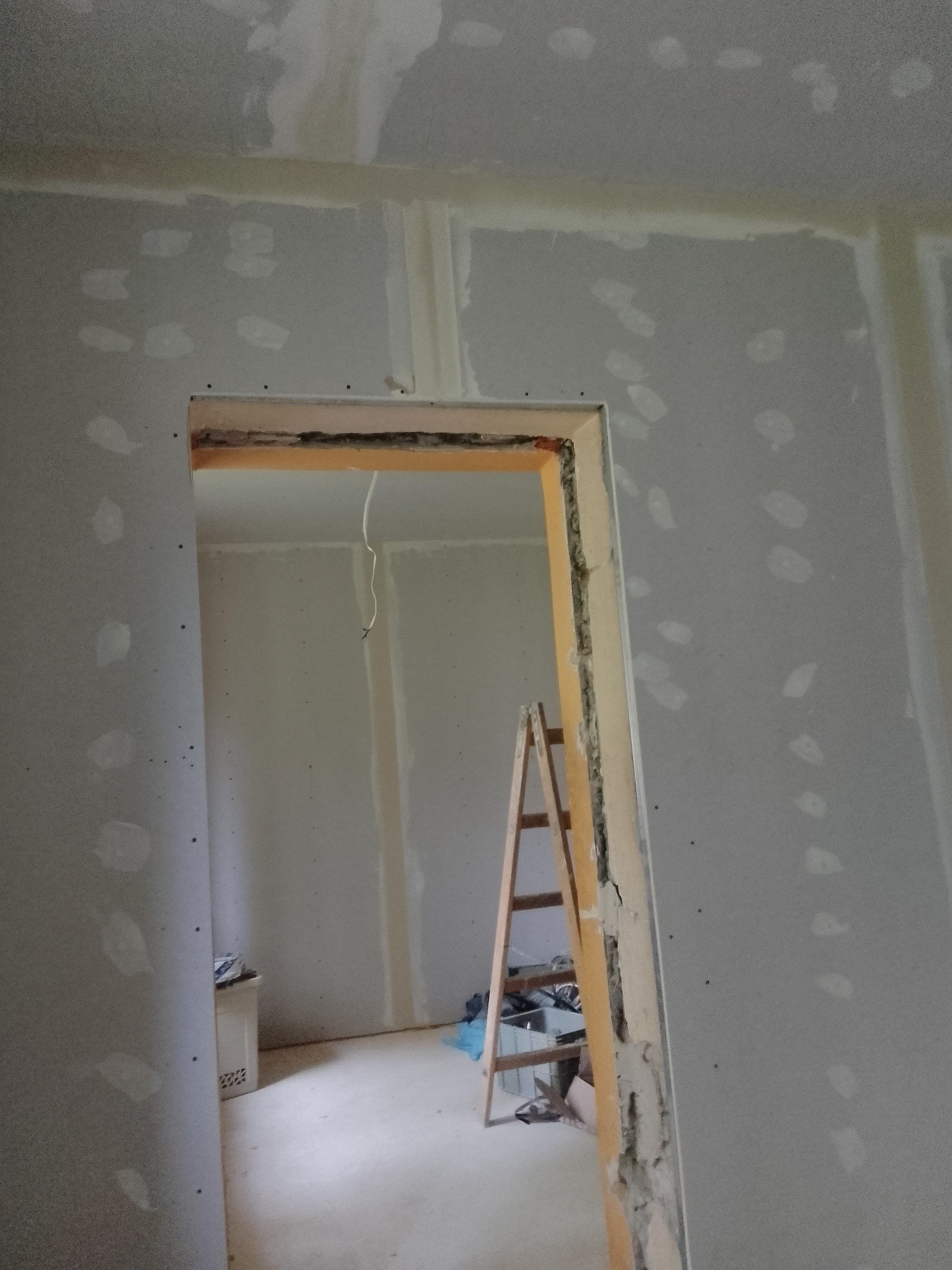 Wnętrze w trakcie prac wykończeniowych: ściany obłożone płytami kartonowo-gipsowymi z widocznymi punktowymi wypełnieniami, drewniana drabina oparta o ścianę, surowa ościeżnica drzwiowa.