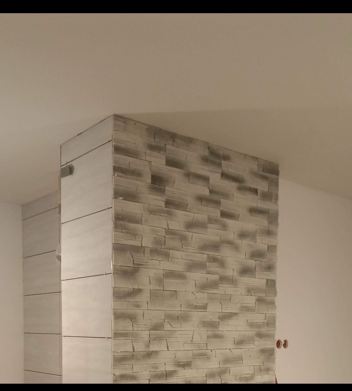 Ściana wykończona dekoracyjną, szarą okładziną imitującą kamień, przylegająca do kolumny obłożonej jasnymi, gładkimi płytkami, widoczne gniazdka elektryczne na sąsiedniej, pomalowanej na biało...