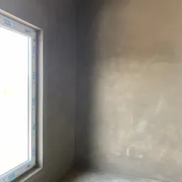 Wnętrze pokoju po nałożeniu tynków maszynowych, widoczna ściana z szarym tynkiem, okno VEKA po lewej stronie, surowy betonowy sufit.