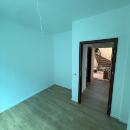 Krystian Kubiczek - Pomalowane na biało wnętrze z drewnianą podłogą i otwartymi drzwiami, przez które widać schody. Z sufitu zwisa kabel. Świeżo po malowaniu.