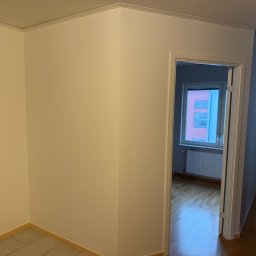 Krystian Kubiczek - Jasne, świeżo pomalowane ściany w pokoju z widocznymi detalami wykończeniowymi, listwami przypodłogowymi i ościeżnicami. Widok na sąsiednie pomieszczenie z oknem.