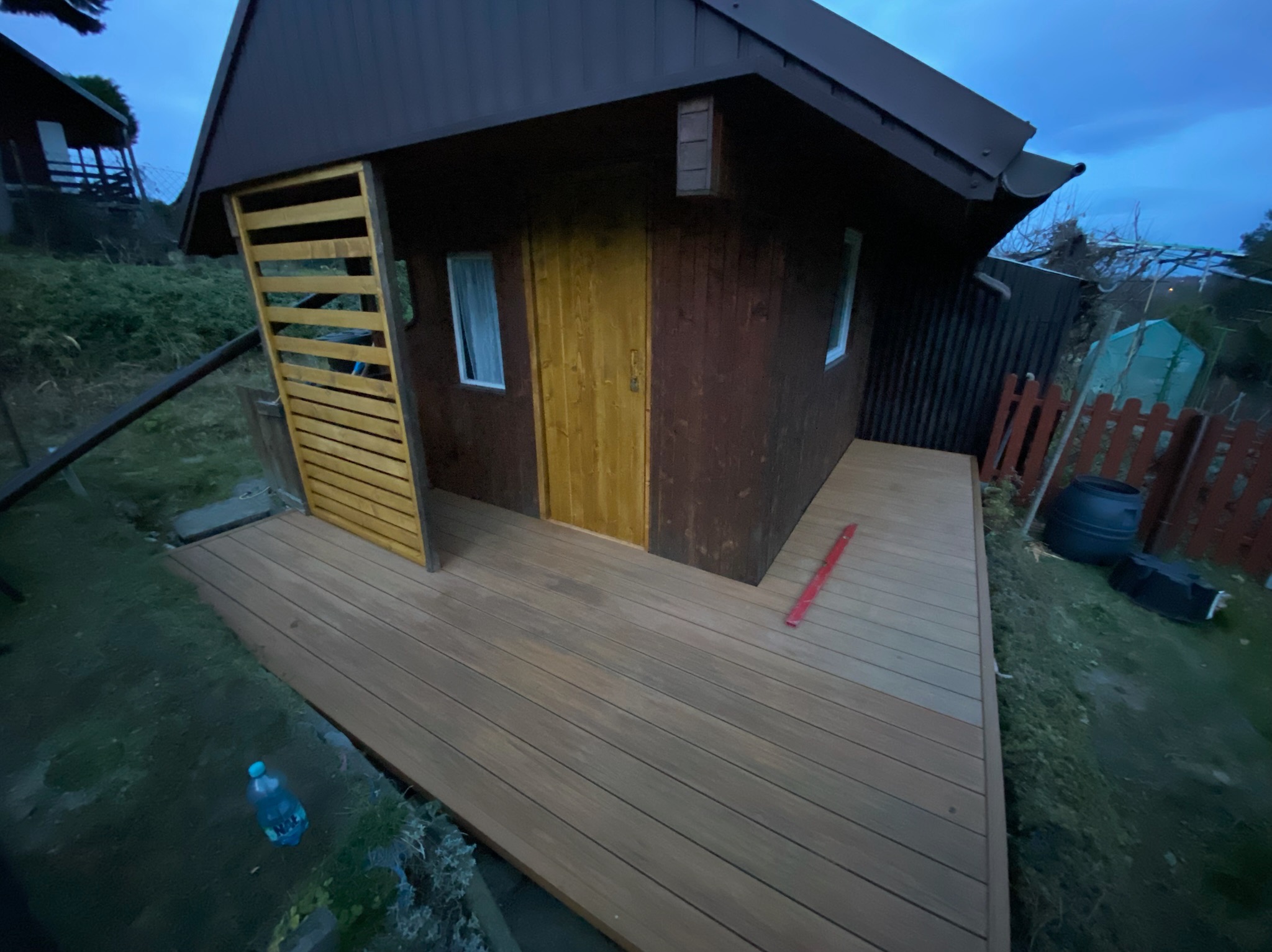 Drewniany domek z nową, brązową deską tarasową. Widoczna butelka wody i poziomica. Ujęcie z dołu, podkreślające przestrzeń tarasu i estetykę wykończenia.