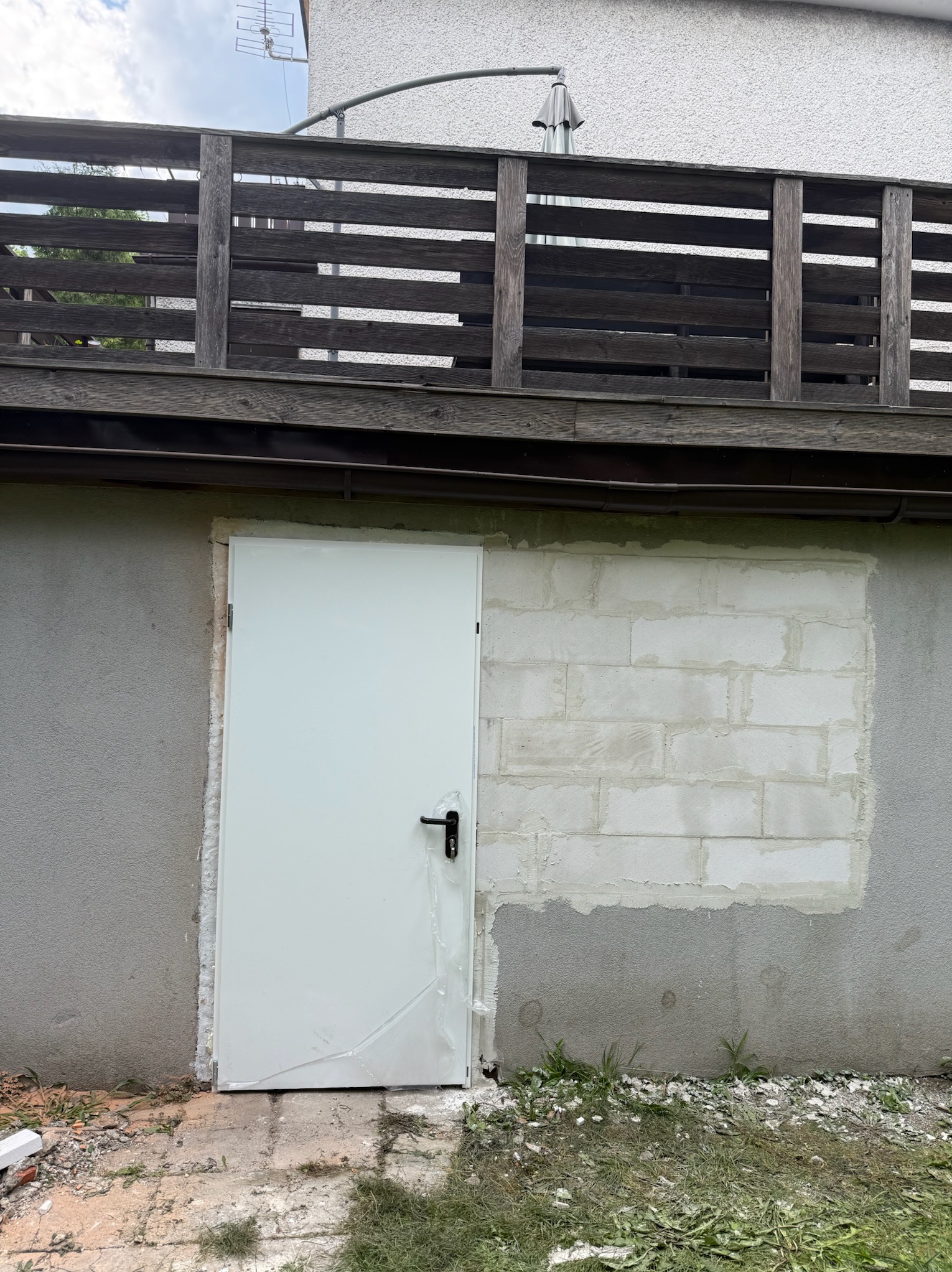 Białe drzwi wejściowe w szarej ścianie budynku z fragmentem zamurowanego otworu z bloków. Widoczny fragment drewnianej balustrady balkonu.