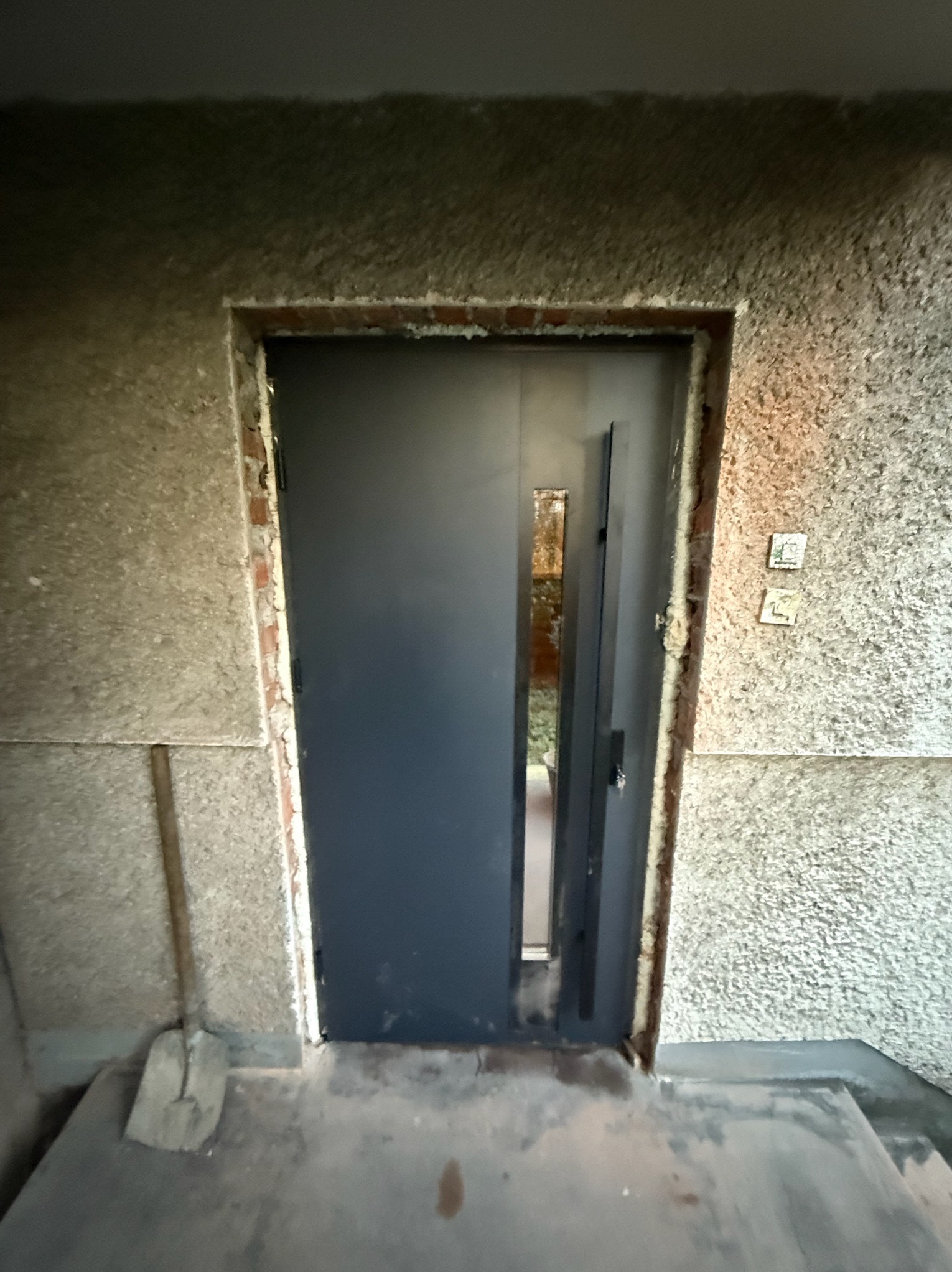 Montaż drzwi wejściowych w Krakowie. Nowoczesne, grafitowe drzwi z wąskim przeszkleniem, osadzone w surowej, teksturowanej ścianie. Widoczne ślady prac montażowych.