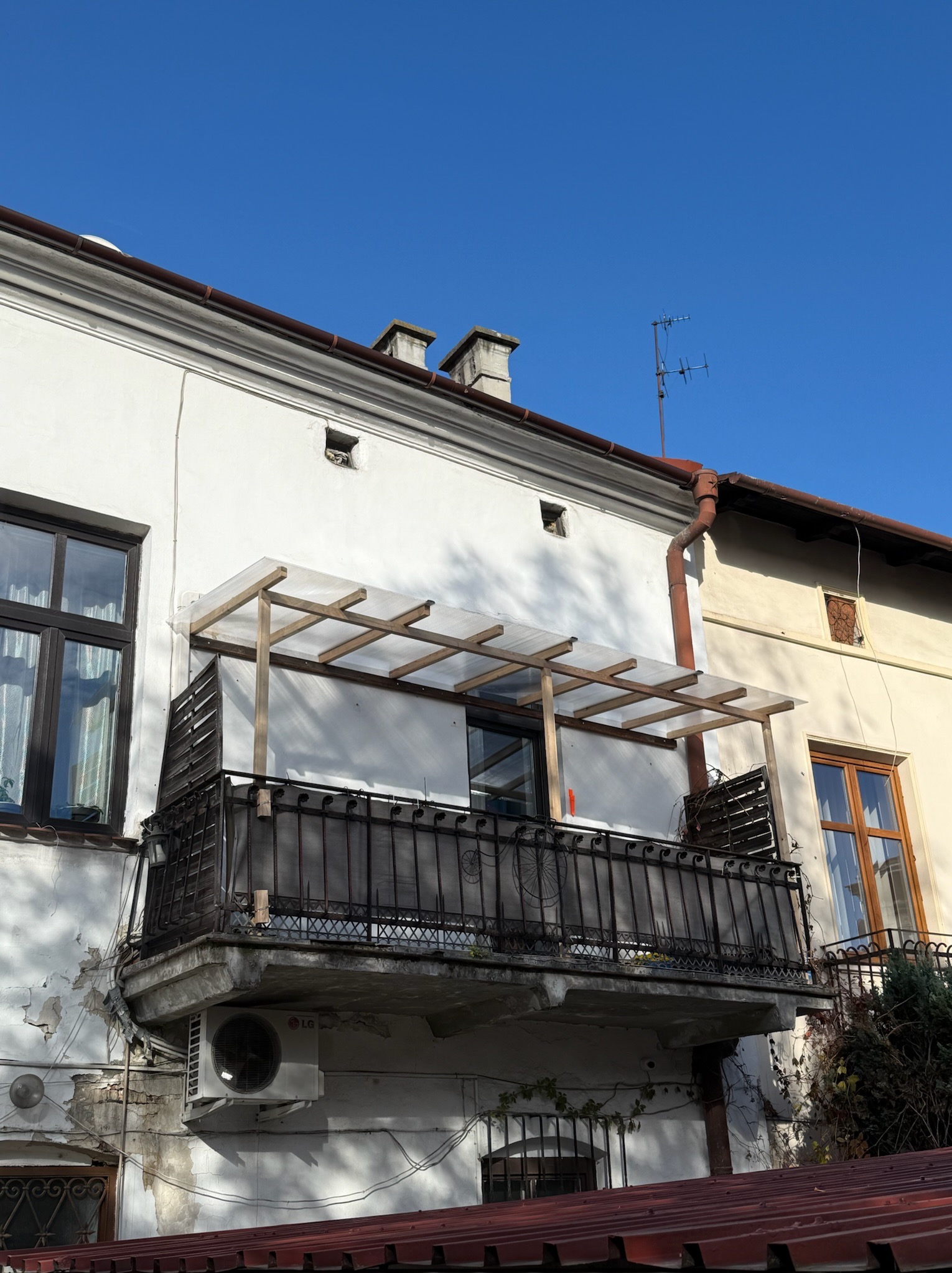 Balkon z drewnianą konstrukcją zadaszenia i metalową balustradą na tle białej, częściowo zniszczonej elewacji budynku w słoneczny dzień.