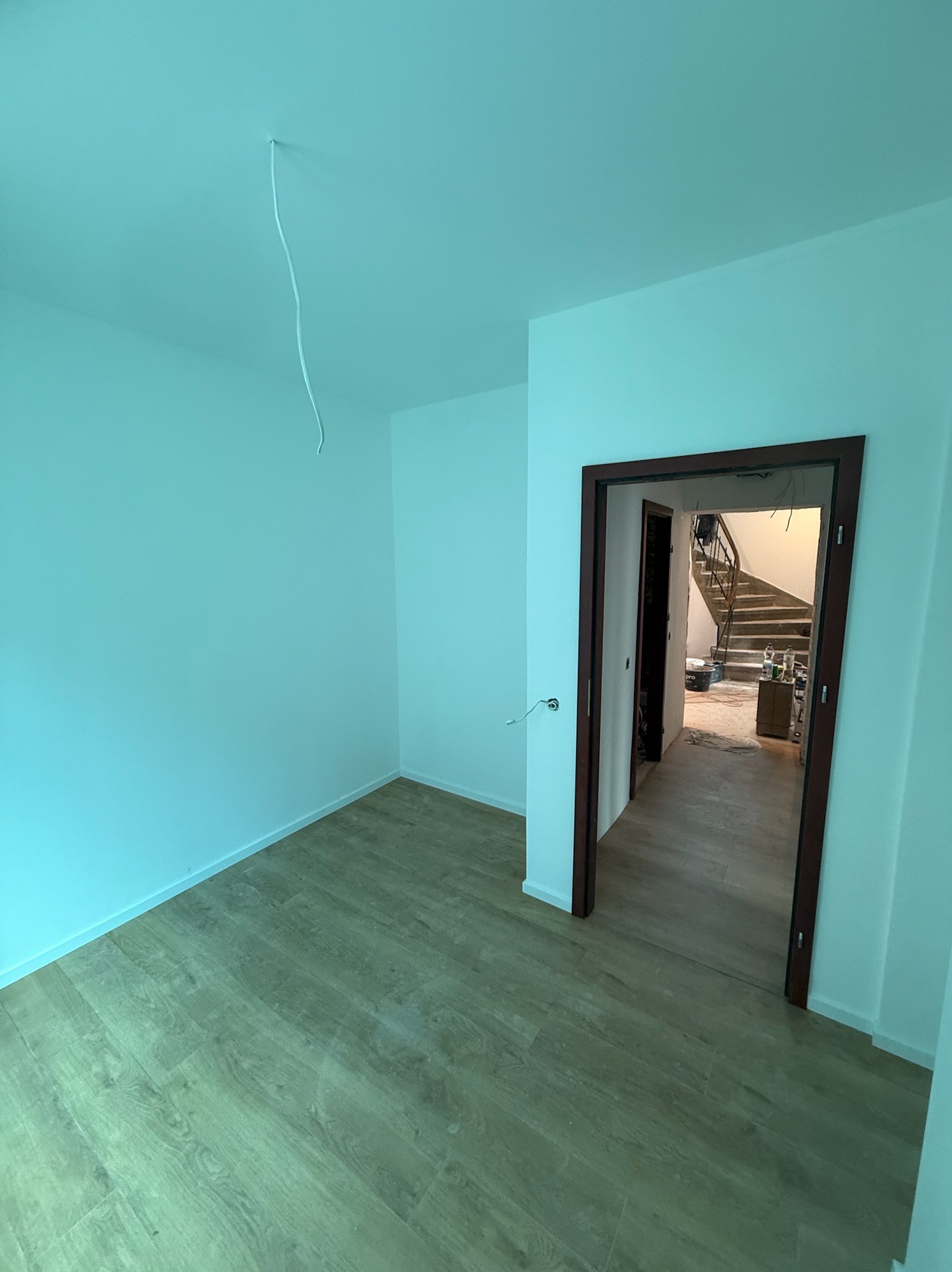 Pomalowane na biało wnętrze z drewnianą podłogą i otwartymi drzwiami, przez które widać schody. Z sufitu zwisa kabel. Świeżo po malowaniu.