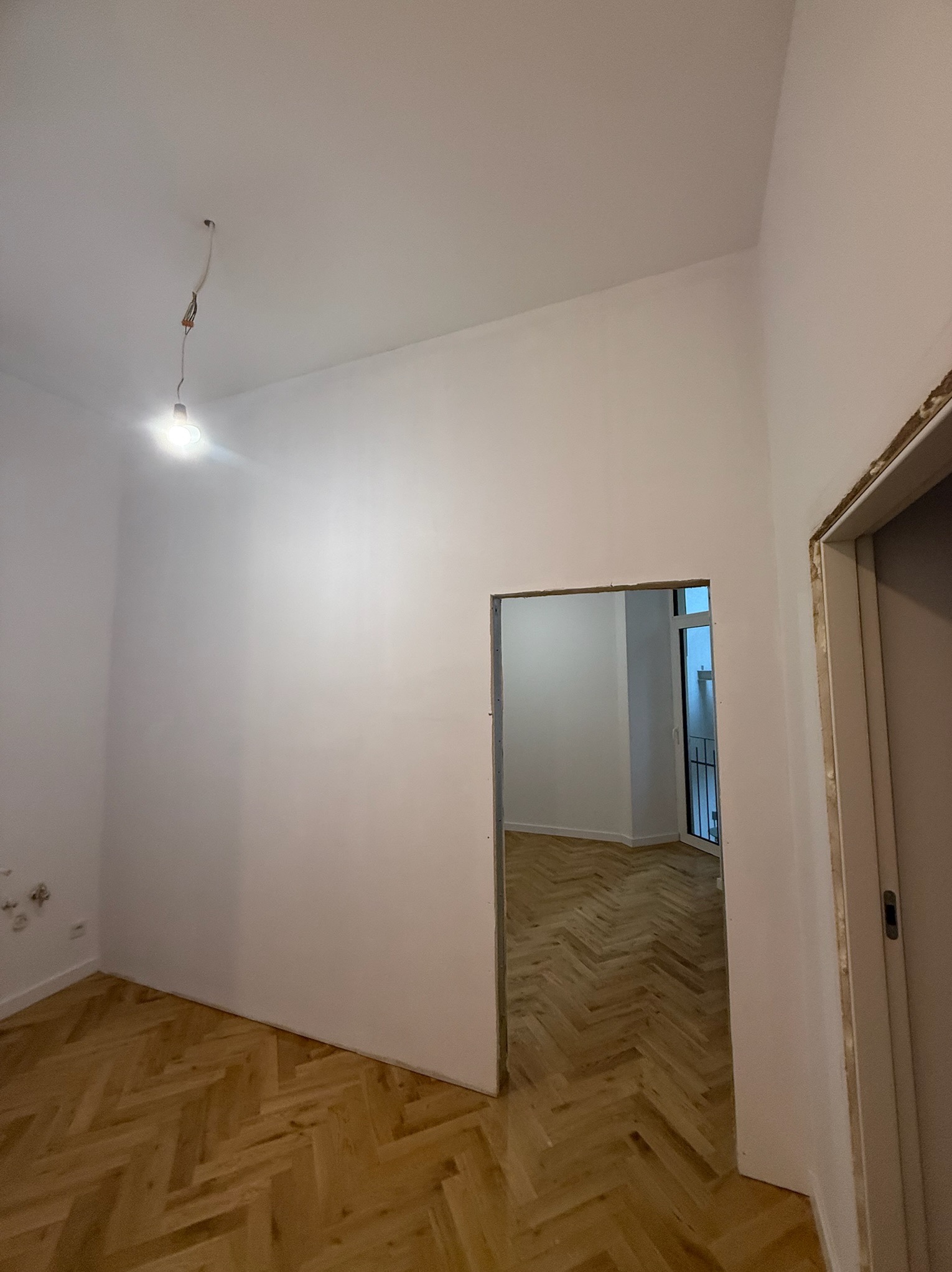 Wnętrze po malowaniu: białe ściany, parkiet w jodełkę, widoczne ślady po gniazdkach i niedokończone obramowanie drzwi. Żarówka na kablu zwisa z sufitu.