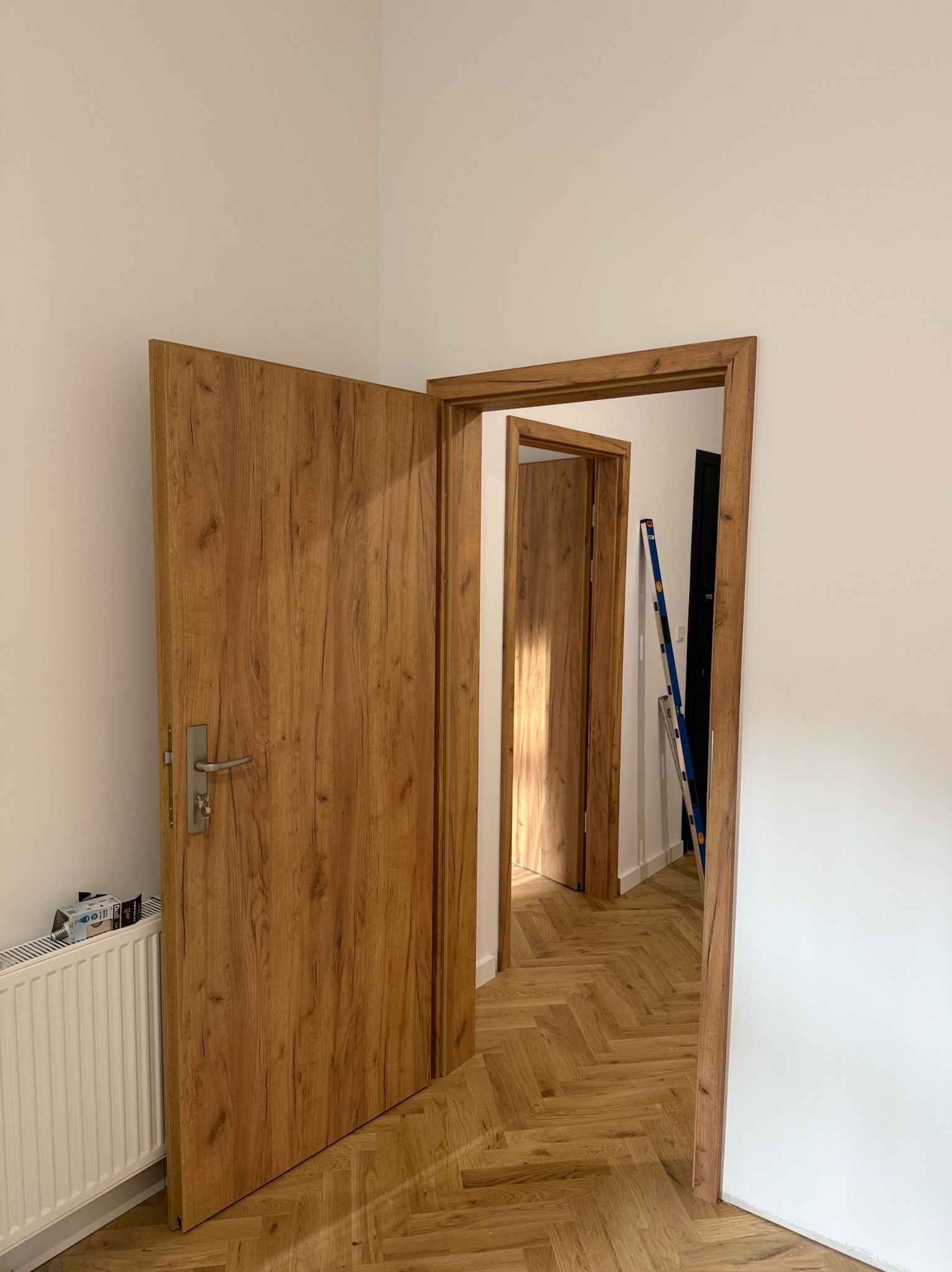 Montaż drzwi wewnętrznych z drewnianą okleiną w nowym mieszkaniu. Widoczny parkiet w jodełkę i białe ściany. W tle drabina oparta o ścianę.