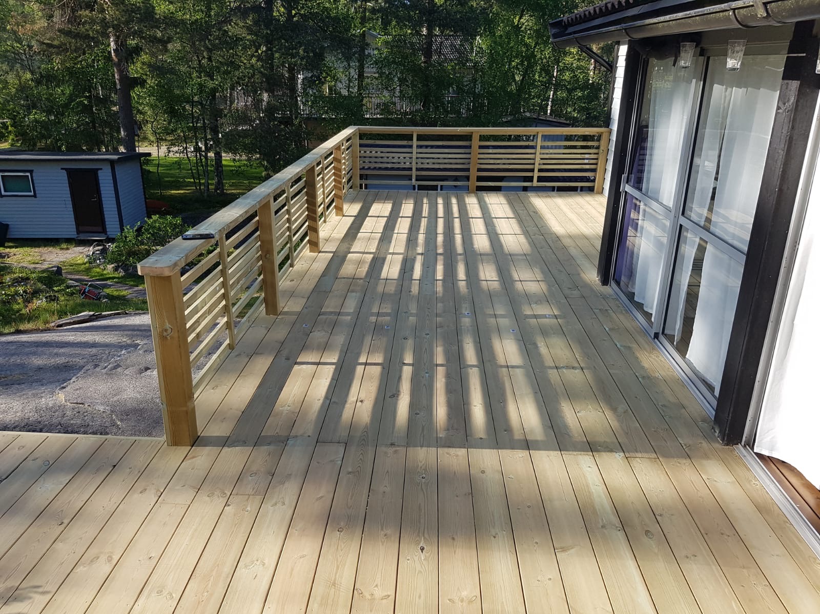 Nowy, drewniany taras z jasnego drewna, z balustradą i widocznymi cieniami, przylegający do budynku z przesuwnymi drzwiami balkonowymi. Widok na zieleń w tle.