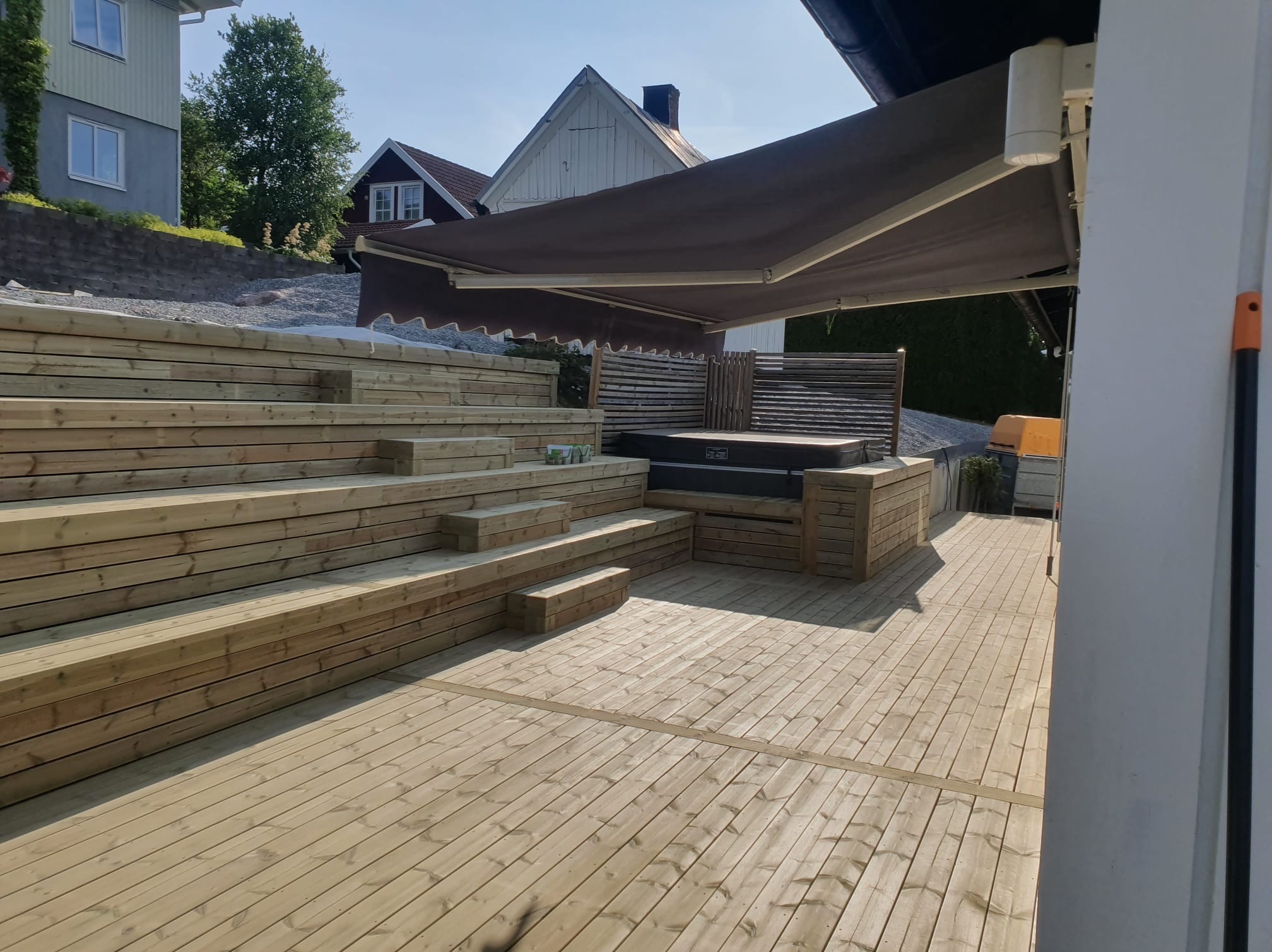 Drewniany taras z jacuzzi i schodami, zadaszony markizą. Jasne drewno, nowoczesny design, dom w tle. Realizacja z dbałością o detale.