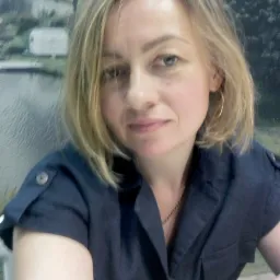 Portret kobiety z blond włosami, w granatowej koszuli z podwiniętymi rękawami, z zegarkiem na brązowym pasku, na tle tapety z motywem jeziora i lasu.