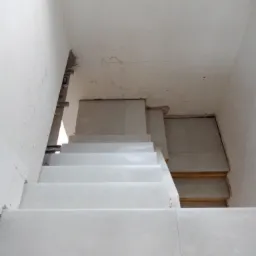 Montaż płytek na schodach wewnętrznych, widoczne niedokończone ściany i drewnopodobna podłoga na dole schodów.