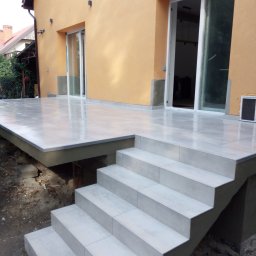 Lubarski Krzysztof - Wykończony taras z jasnoszarych płytek ceramicznych, prowadzący schodami do budynku o pomarańczowej elewacji. Widoczne niedokończone prace ziemne wokół konstrukcji tarasu.