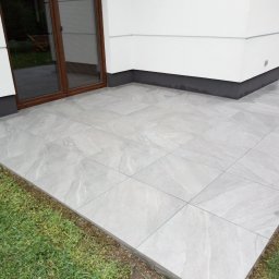 Lubarski Krzysztof - Jasnoszary taras z płytek ceramicznych, widok z góry, z brązowymi drzwiami balkonowymi i fragmentem trawnika. Minimalistyczny design.