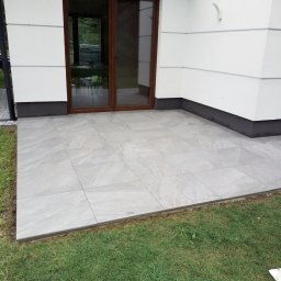 Lubarski Krzysztof - Jasnoszary taras z płytek ceramicznych przy białym budynku z brązowymi drzwiami przesuwnymi. Widok na trawnik. Minimalistyczny design.