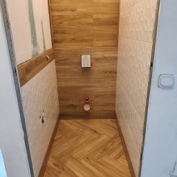 Lubarski Krzysztof - Wnętrze łazienki w trakcie wykańczania: drewnopodobne płytki na ścianach i podłodze ułożone w jodełkę, białe płytki 3D na ścianie, widoczne elementy instalacji.