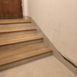 Lubarski Krzysztof - Wykończone schody z drewnopodobnymi stopniami i jasnymi płytkami na podłodze i ścianach. Widoczne ślady po pracach wykończeniowych na ścianie.