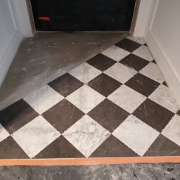 Lubarski Krzysztof - Układanie podłogi z płytek w szachownicę, widoczne krzyżyki dystansowe, część podłogi jeszcze nie wykończona, zabezpieczone drzwi w tle.