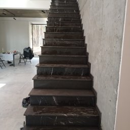 Lubarski Krzysztof - Schody wewnętrzne wyłożone ciemnym kamieniem z białymi żyłami, prowadzące w górę, widoczne z dołu, surowa ściana z betonu architektonicznego obok, w tle widoczny stół i krzesła.