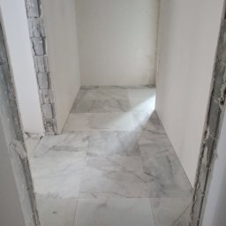 Lubarski Krzysztof - Wnętrze w trakcie prac wykończeniowych: ułożone marmurowe płytki podłogowe, surowe ościeżnice drzwiowe z widocznymi śladami zaprawy, świeżo pomalowane ściany.
