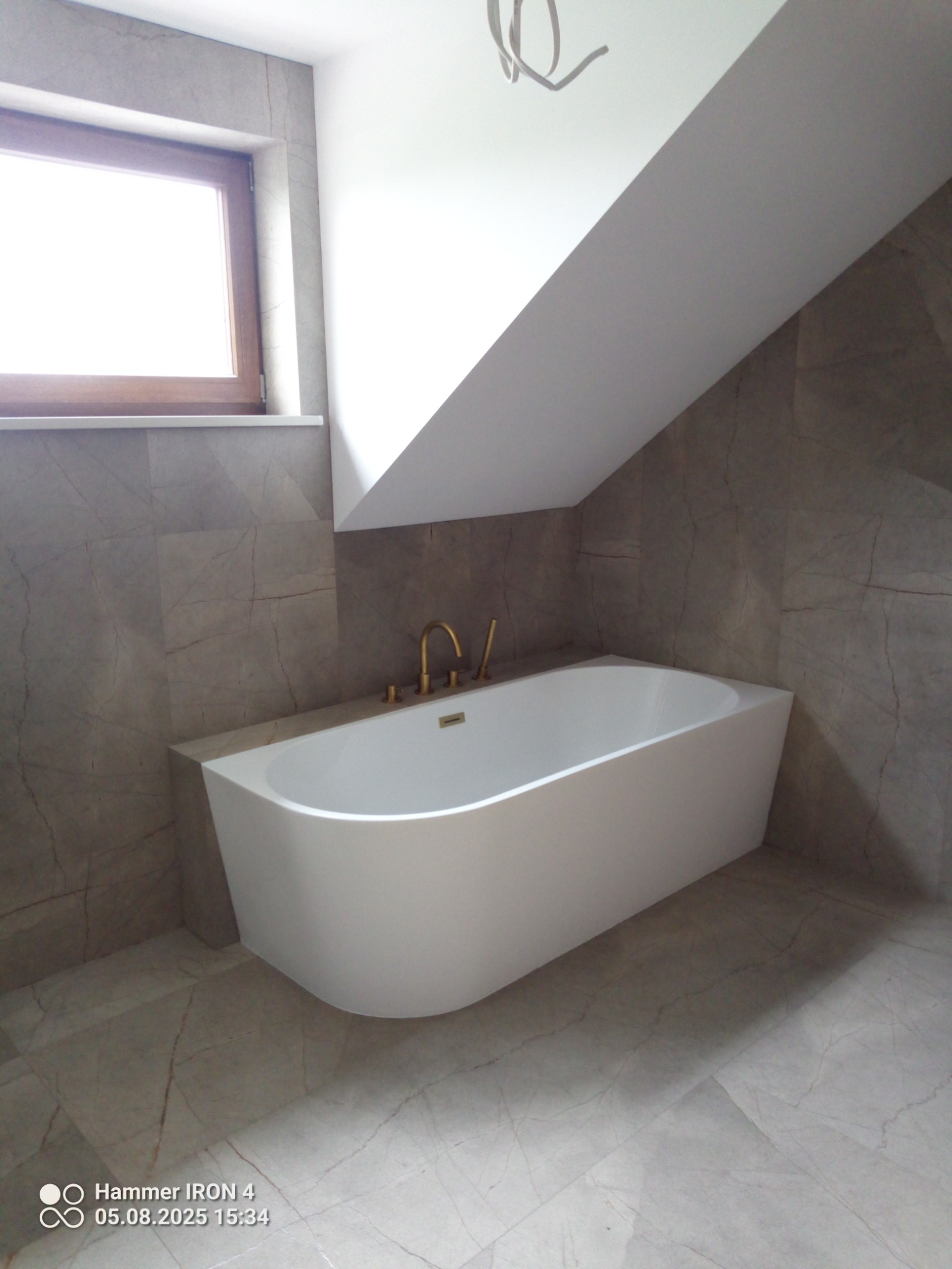 Nowoczesna, wolnostojąca wanna w łazience wykończonej płytkami imitującymi marmur. Złota armatura, okno w tle. Ukośny sufit dodaje charakteru wnętrzu.