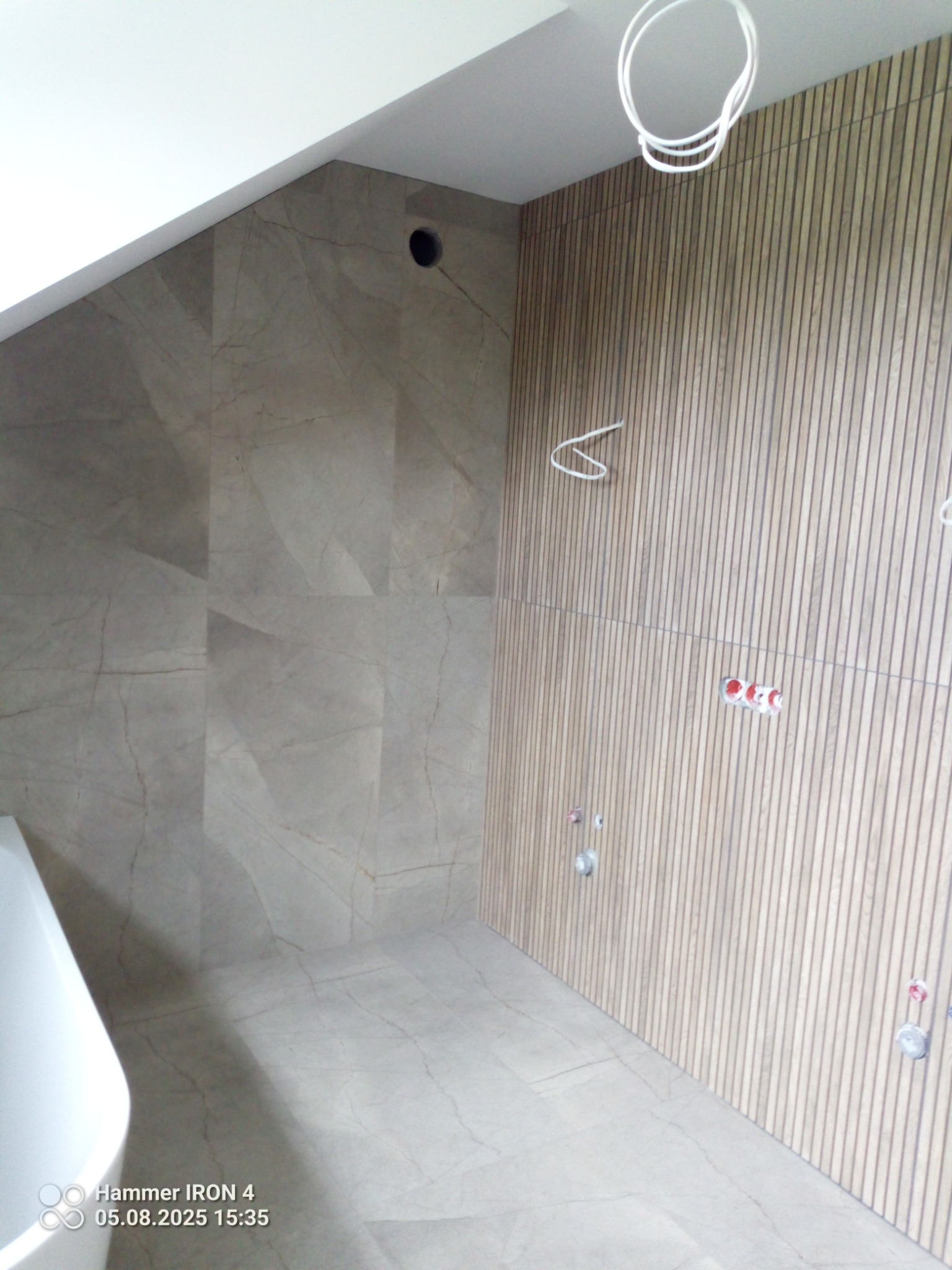 Wykończona łazienka: płytki imitujące marmur na ścianie, drewniane panele pionowe. Widoczne instalacje. Częściowo widoczna wanna. Jasna kolorystyka.