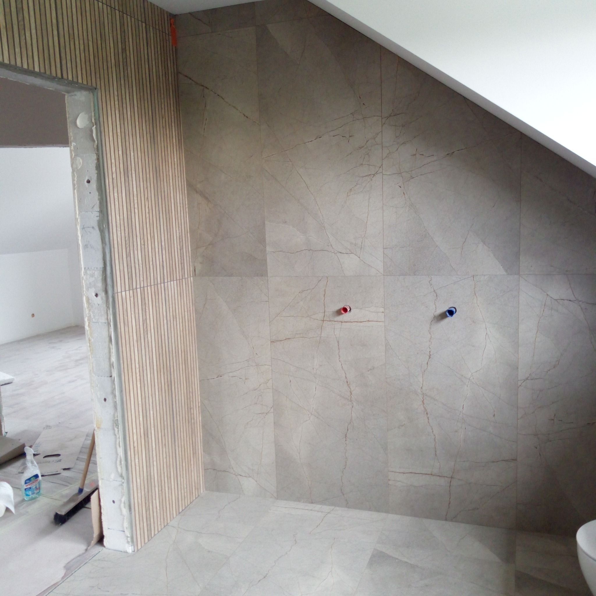 Wykończona łazienka na poddaszu: marmurowe płytki na ścianach i podłodze, drewniane lamele przy wejściu, widoczne przyłącza wodne, ukośny sufit.