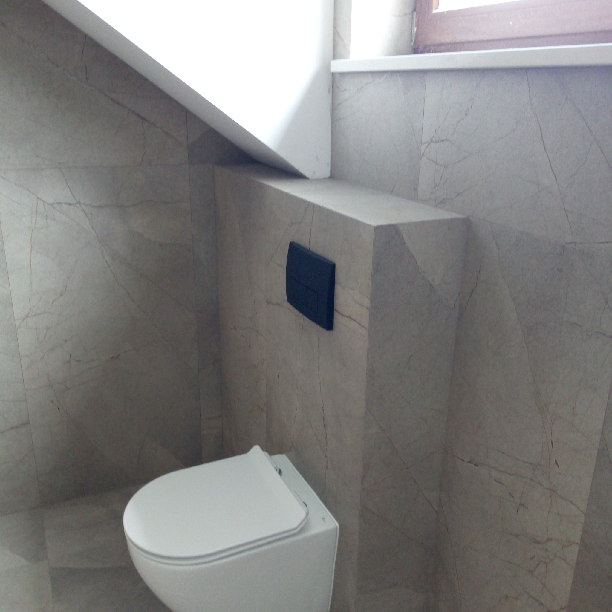 Podwieszana toaleta z czarnym przyciskiem spłukiwania na tle ściany wyłożonej marmurowymi płytami w odcieniach szarości i beżu. Minimalistyczny design wnętrza.