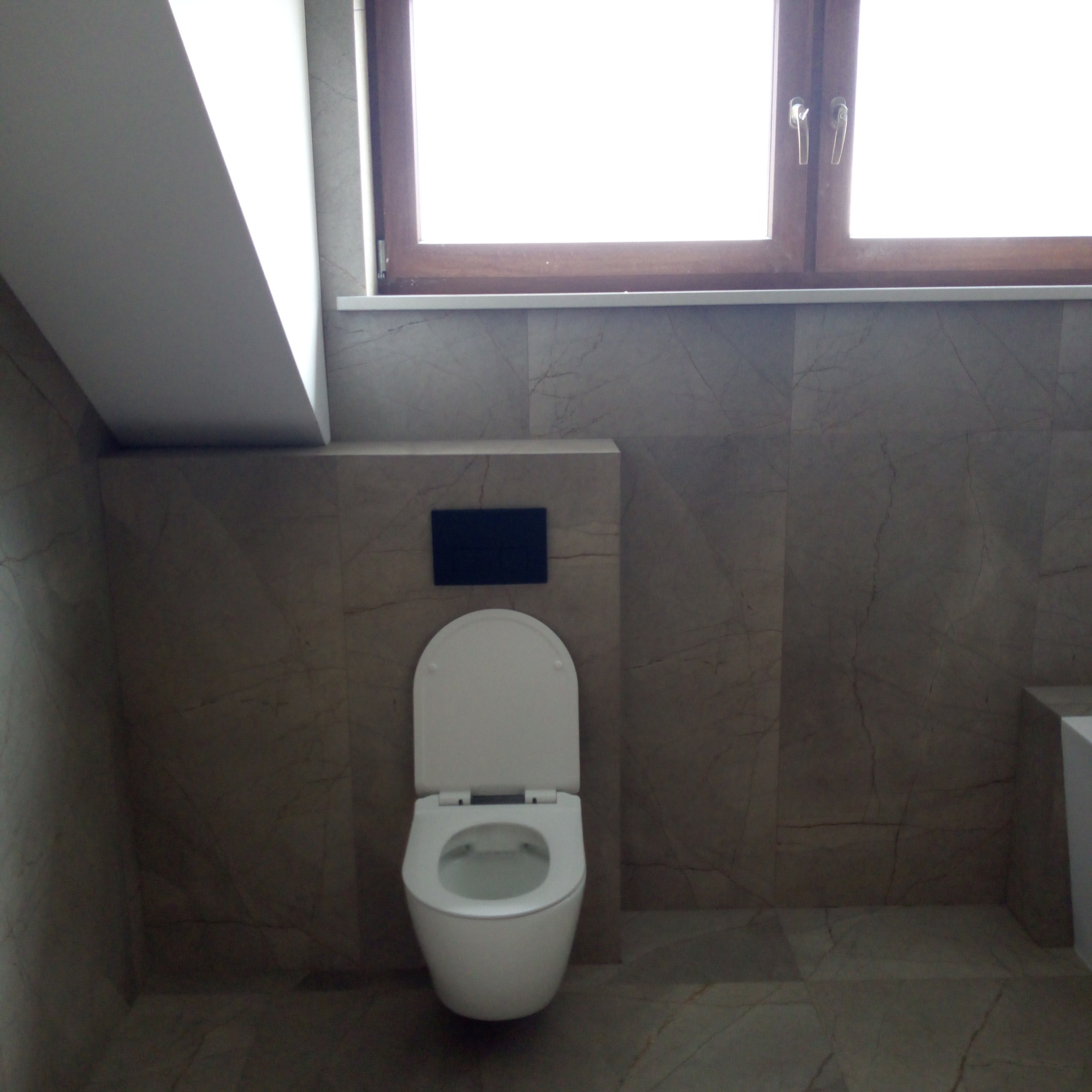 Elegancka łazienka z marmurowymi płytkami i podwieszaną toaletą. Okno z brązową ramą w tle. Ukośny sufit dodaje przestrzeni. Stonowana kolorystyka.