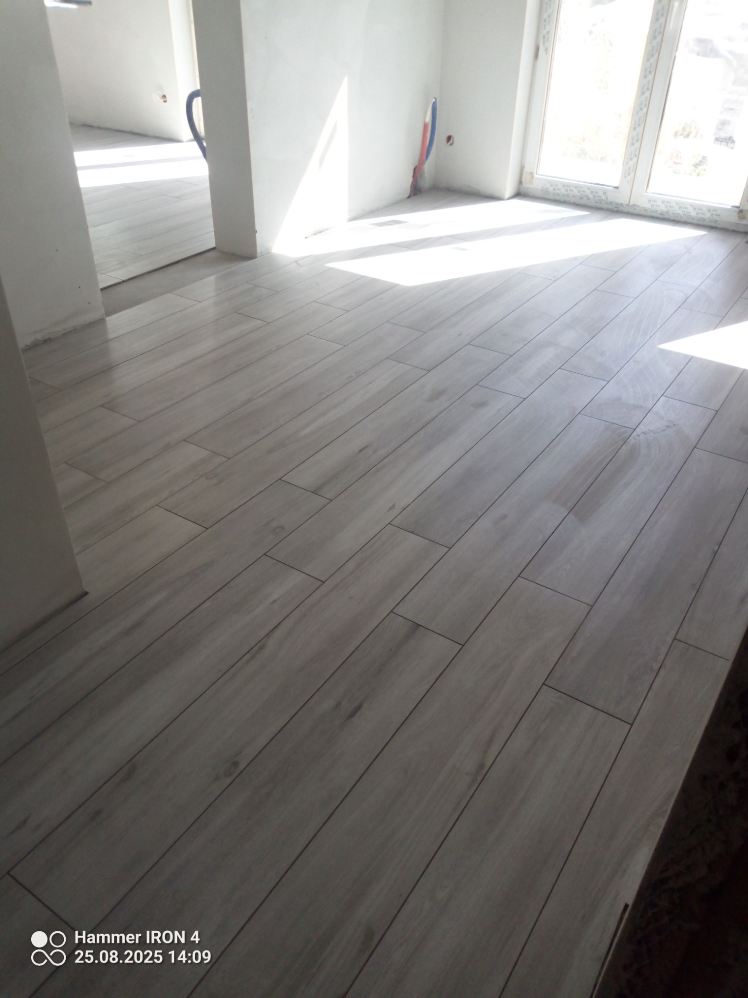 Jasne, drewnopodobne płytki ułożone w jodełkę na podłodze w nowym, jasnym pomieszczeniu. Widoczne fragmenty ścian i okno w tle. Minimalistyczny design.