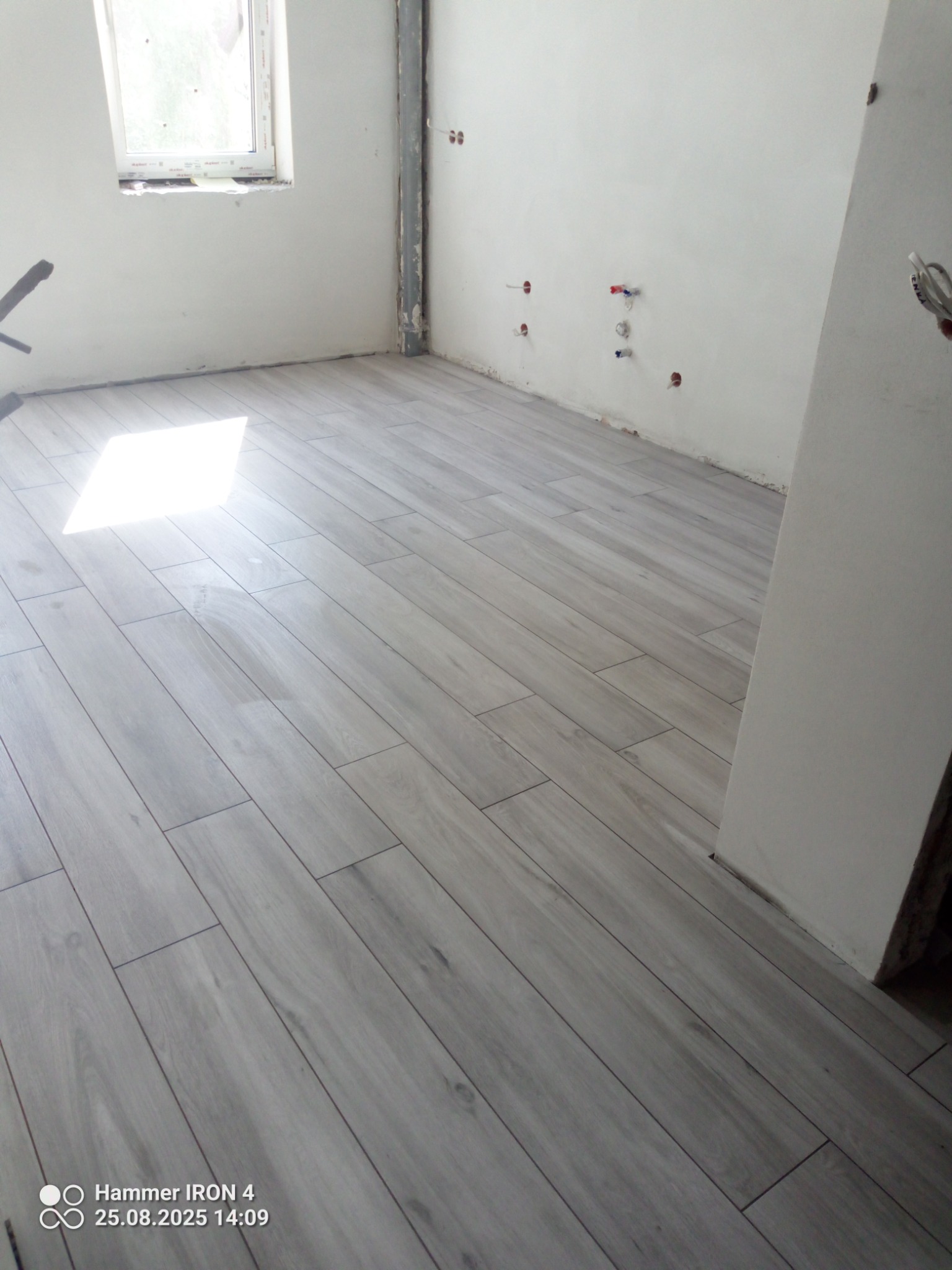 Ułożona, jasna podłoga z płytek imitujących drewno w surowym wnętrzu z oknem i wyprowadzonymi instalacjami. Widoczne połączenia płytek, fuga w kolorze szarym.