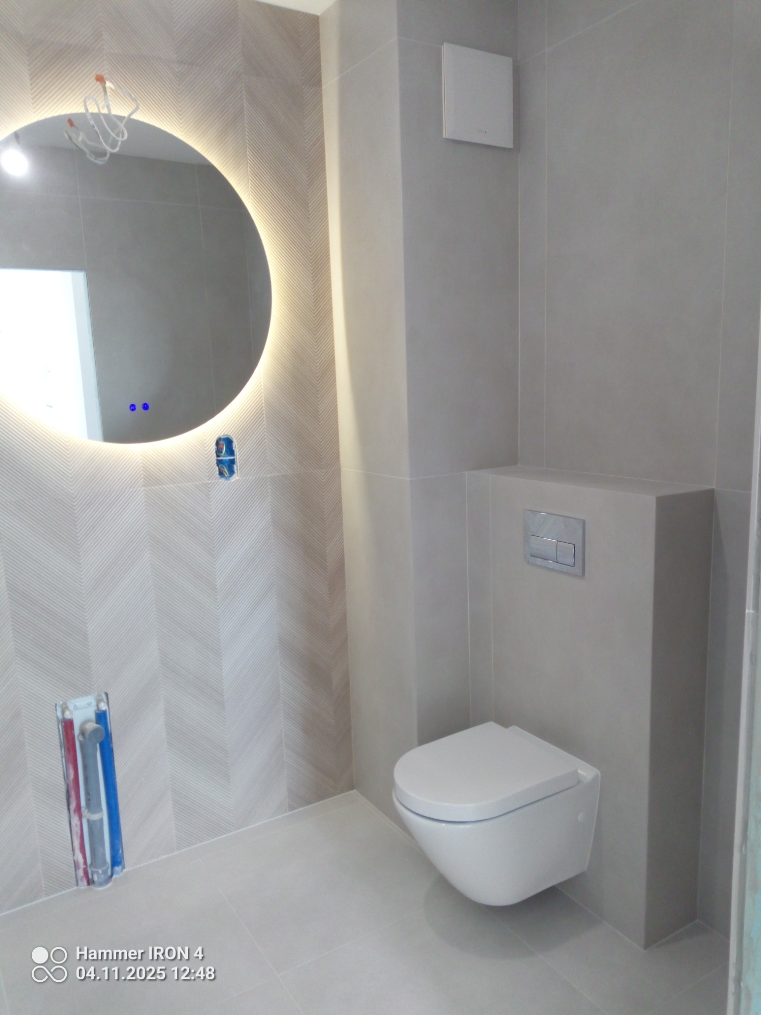 Nowoczesna łazienka z podwieszaną toaletą i okrągłym lustrem z podświetleniem LED. Na ścianie płytki w subtelny wzór jodełki. Minimalistyczny design.