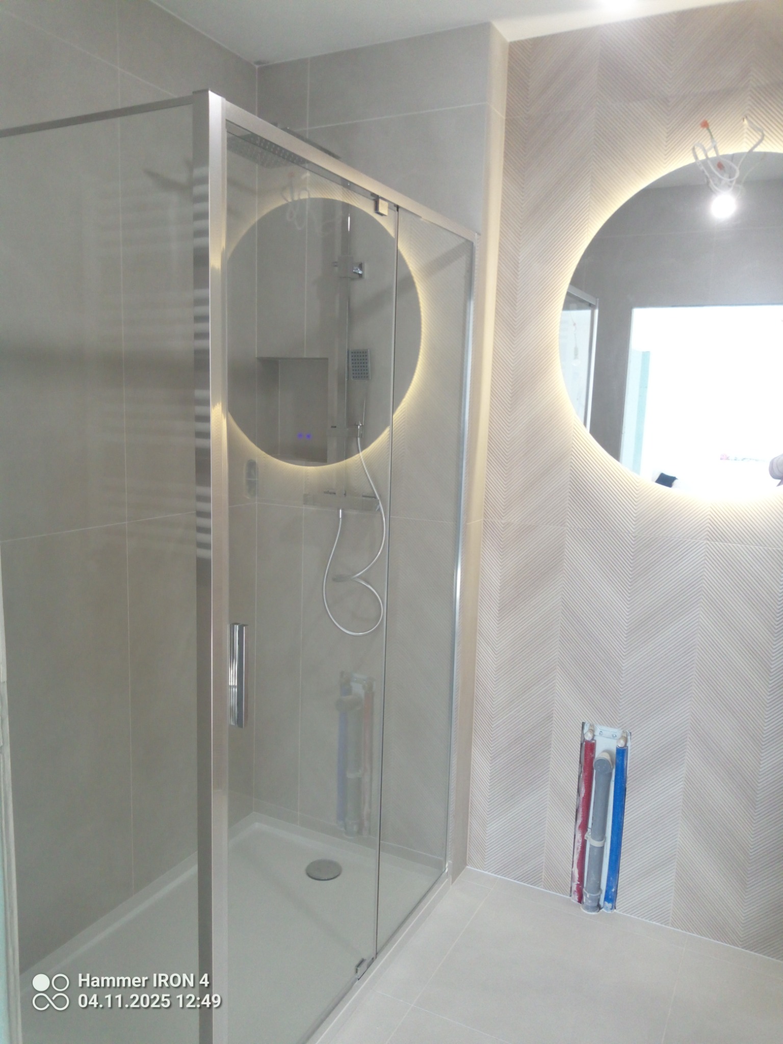 Nowoczesna kabina prysznicowa z okrągłym lustrem podświetlanym LED, jasne płytki z subtelnym wzorem jodełki na ścianie. Minimalistyczny design, widoczne przyłącza wodne.