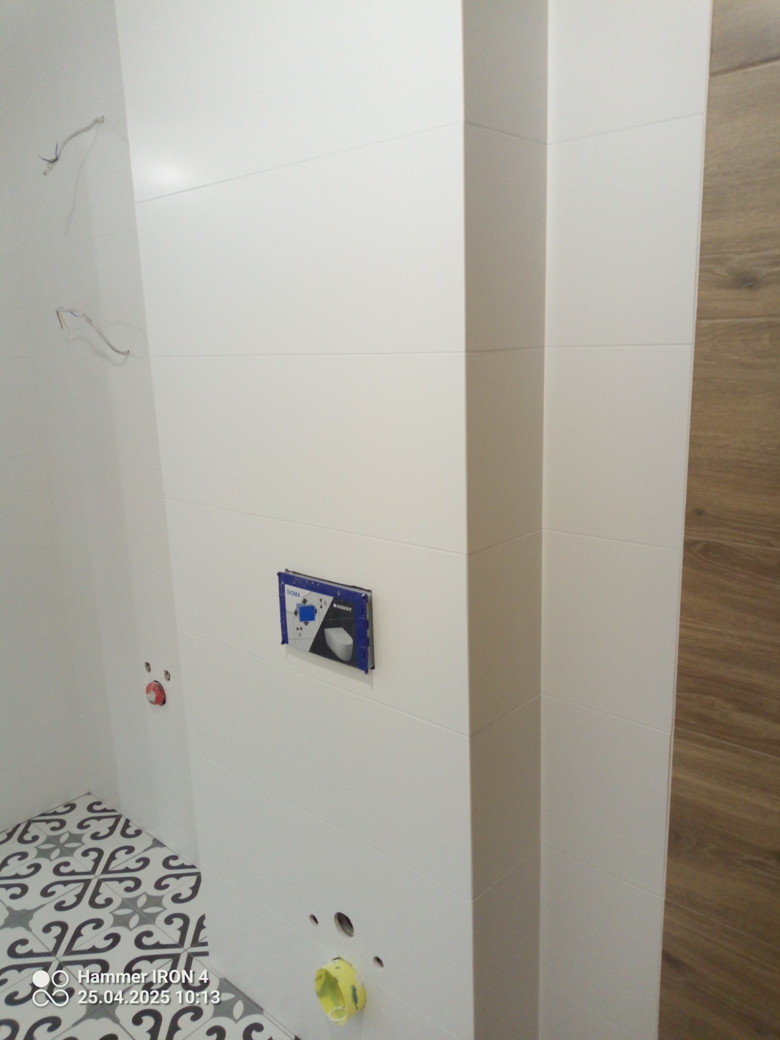 Białe, poziome płytki w łazience, widoczna instalacja podtynkowa WC i fragment podłogi z wzorzystymi płytkami. Częściowo wykończona ściana z drewnopodobnymi panelami.