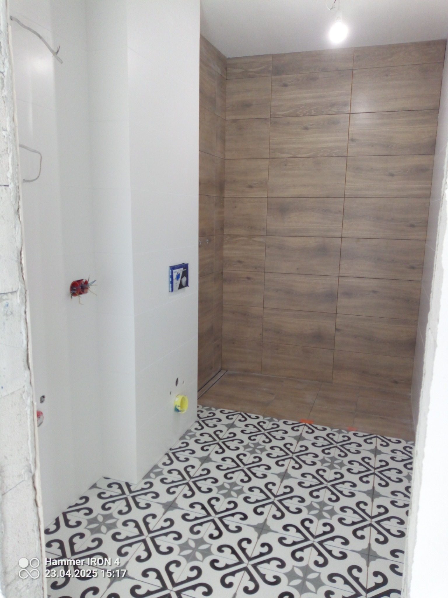 Wykończona łazienka z drewnopodobnymi płytkami na ścianach i wzorzystą podłogą. Widoczne białe ściany z instalacją. Ujęcie z niskiego kąta.