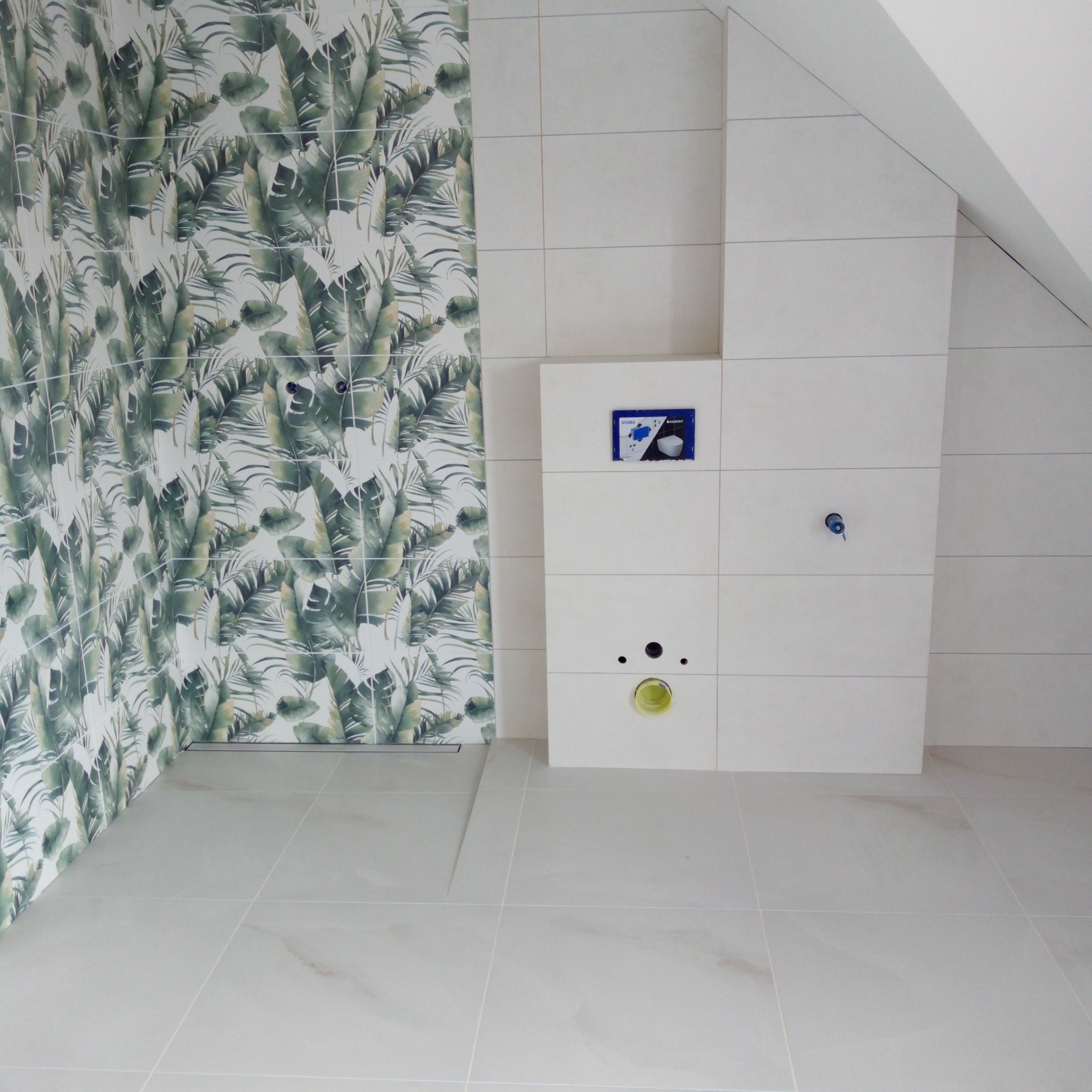 Wykończona łazienka w jasnych płytkach z motywem roślinnym na ścianie. Widoczny stelaż podtynkowy WC i odpływ liniowy. Ukośny sufit.