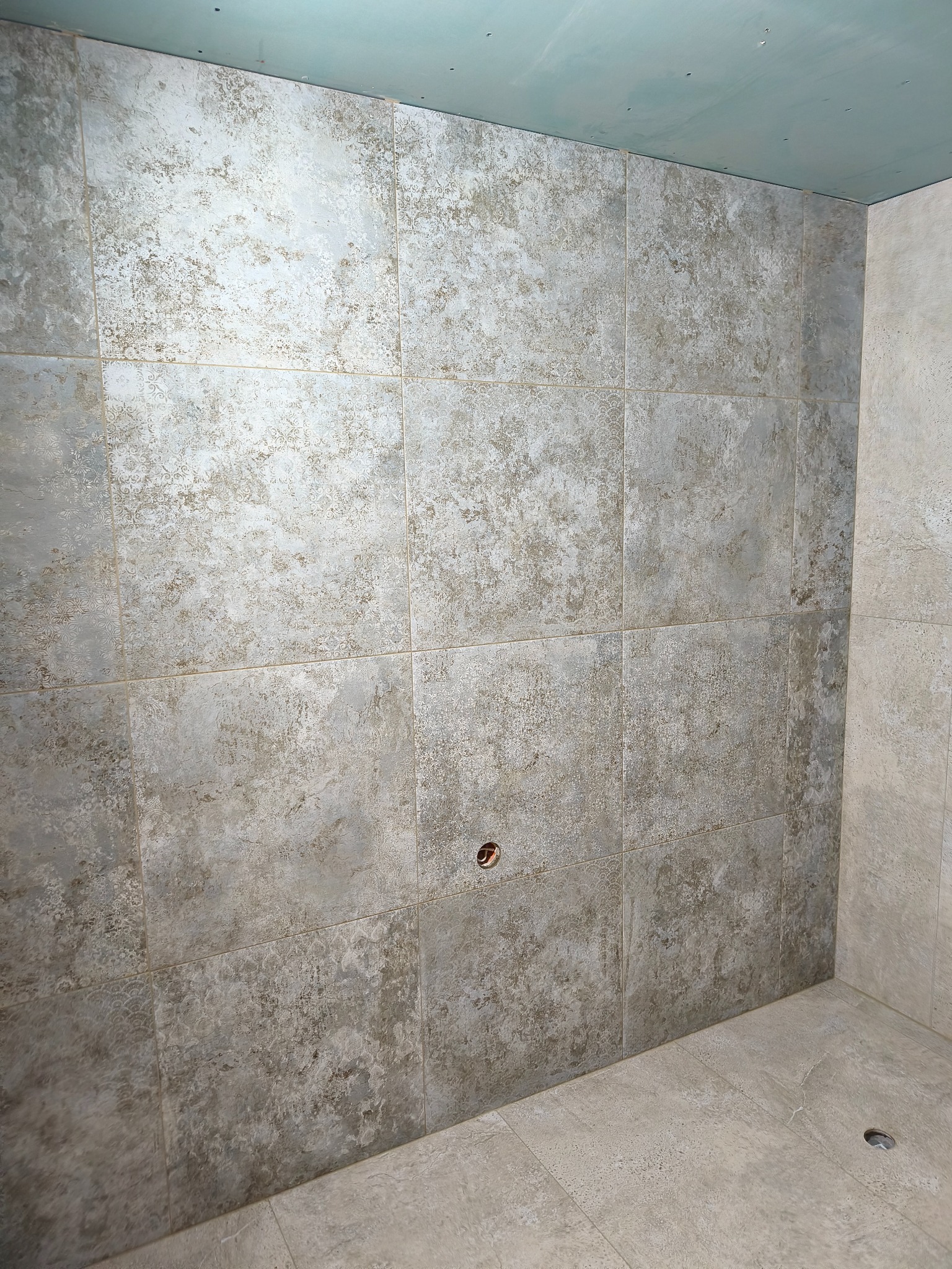 Wykończona ściana w łazience z szarych, wzorzystych płytek ceramicznych. Widoczny fragment podłogi i sufitu. Otwór na instalację w ścianie.