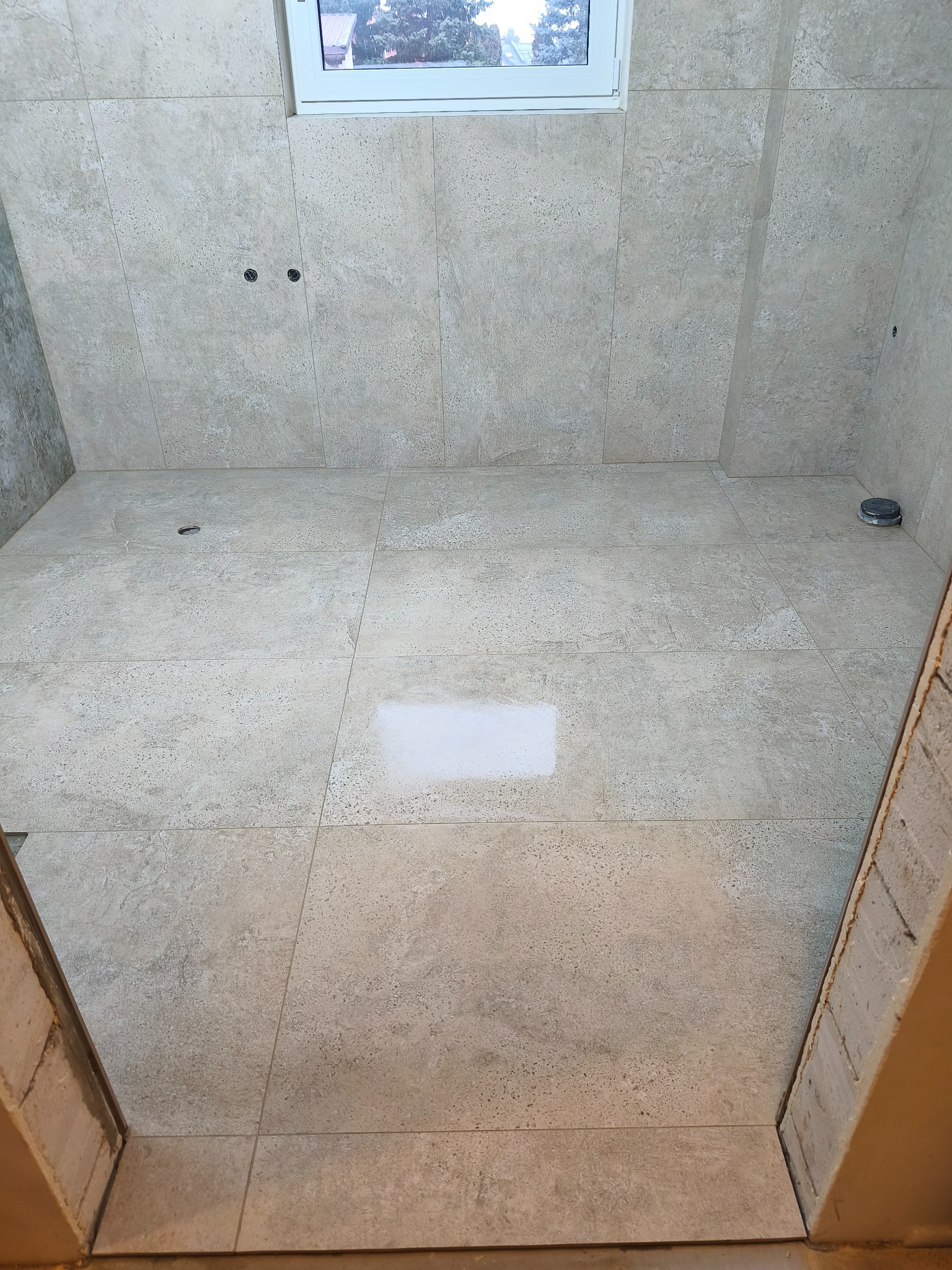 Wykończona łazienka z beżowymi płytkami na ścianach i podłodze. Widoczne otwory na armaturę i okno w górnej części kadru. Ujęcie z dołu, podkreślające przestrzeń.