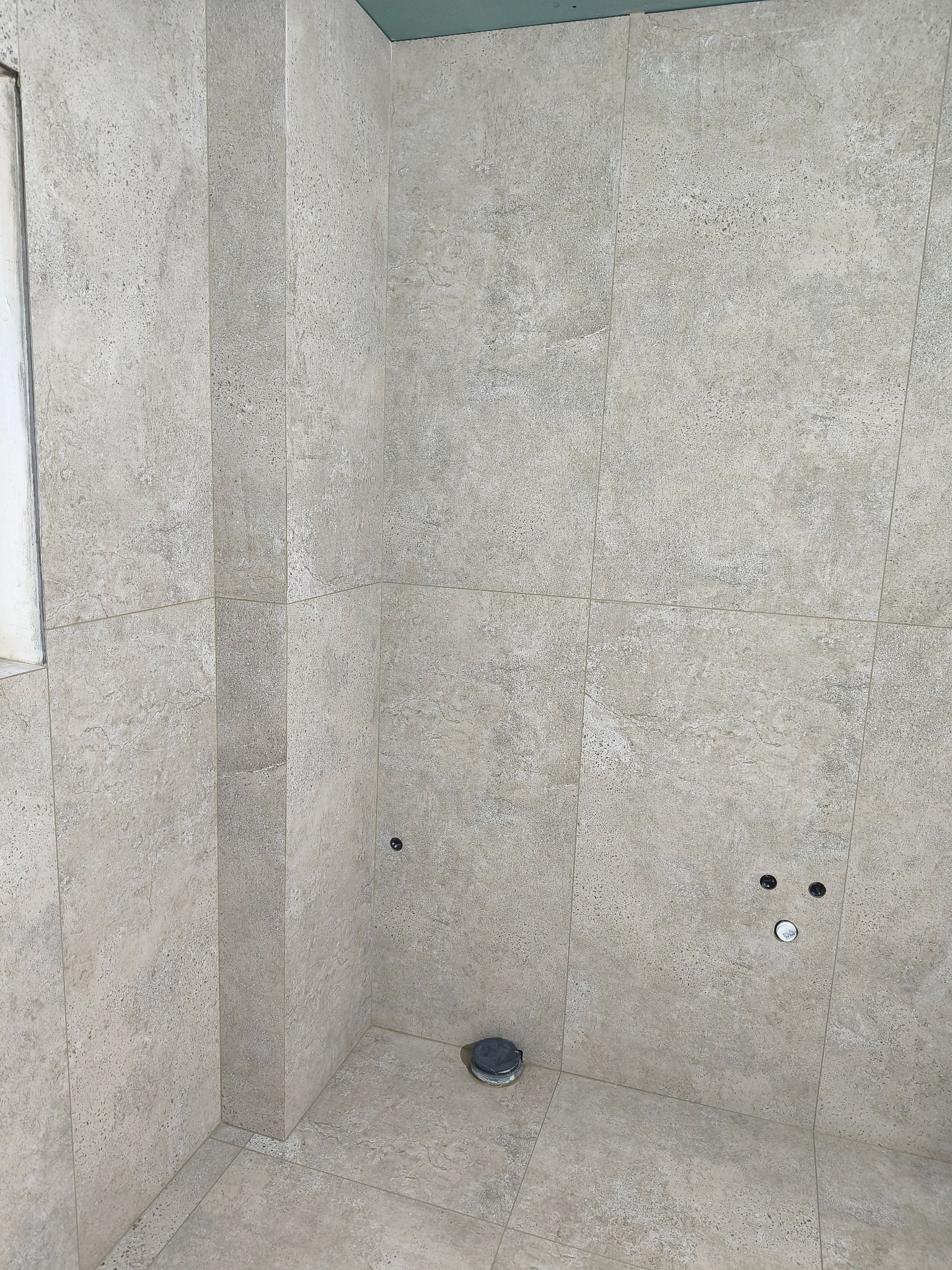 Wykończona w beżowych płytkach kabina prysznicowa z widocznymi przyłączami wodnymi i odpływem podłogowym. Minimalistyczny design, neutralna kolorystyka.