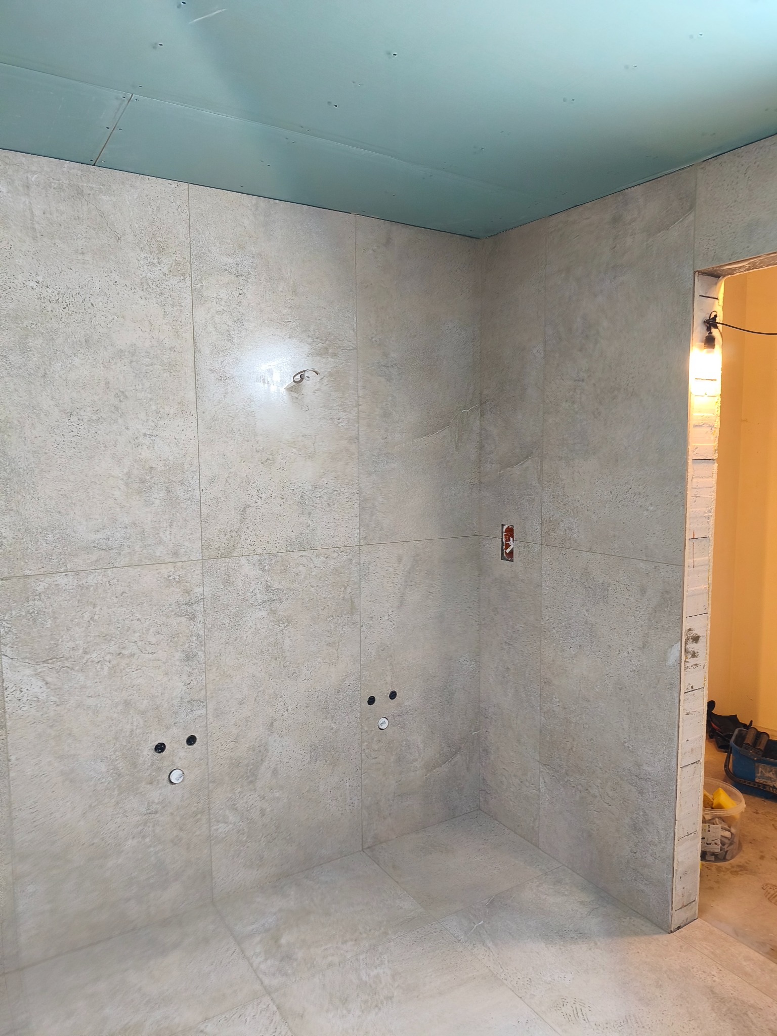 Wykończona beżowa łazienka z płytkami imitującymi kamień, widoczne punkty instalacji wodnej i elektrycznej. Sufit podwieszany z płyt kartonowo-gipsowych.