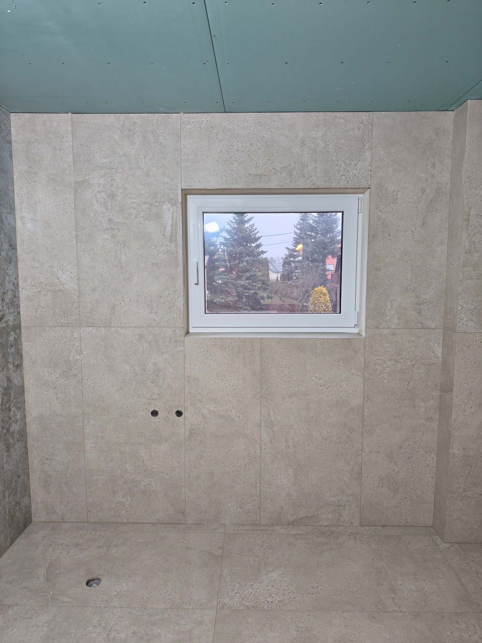 Wykończona beżowa łazienka z oknem, widoczne płytki na ścianach i podłodze, sufit w trakcie wykańczania. Dwa otwory na baterię. Widok z okna na zewnątrz.