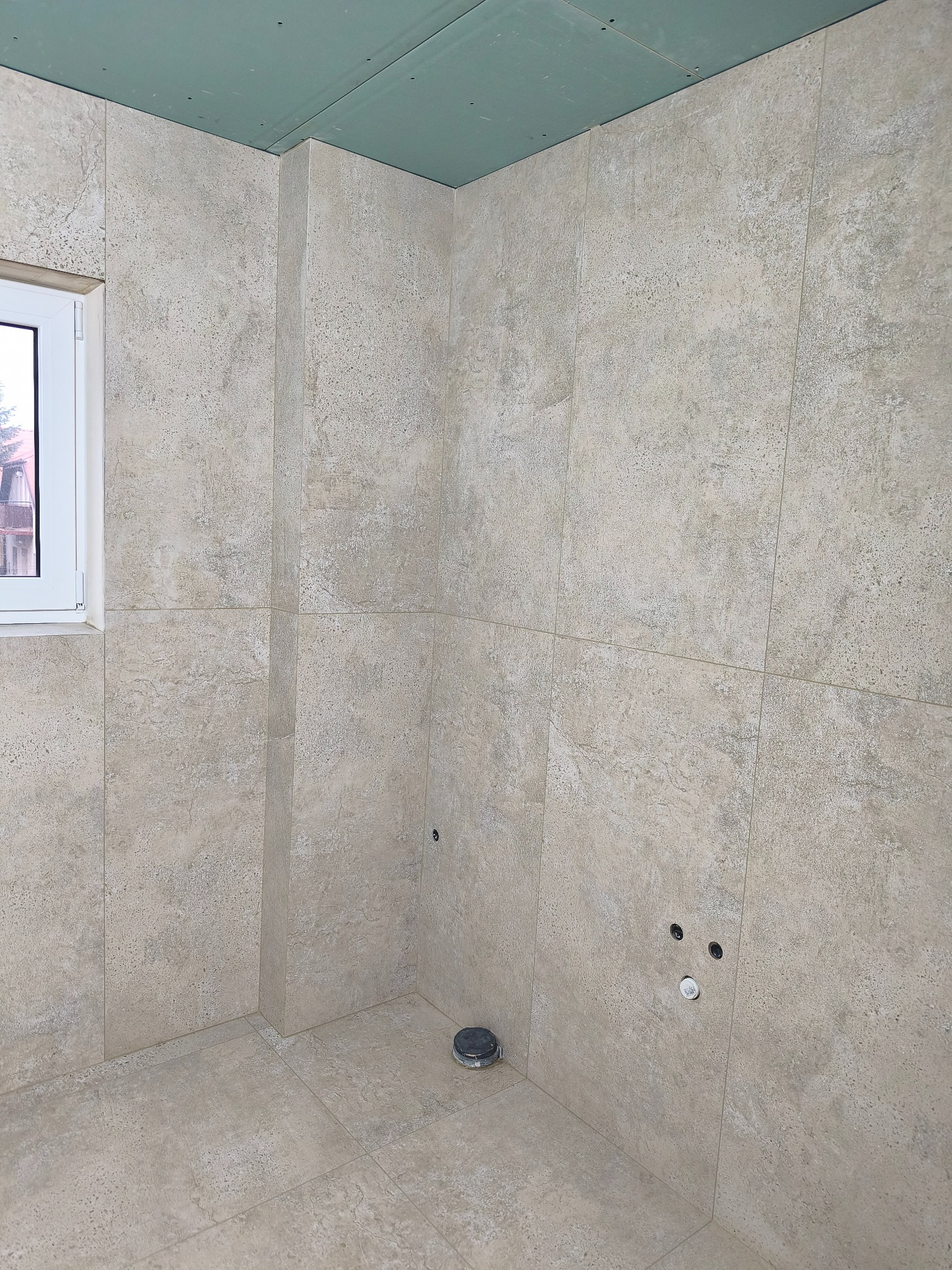 Wykończona łazienka z beżowymi płytkami imitującymi kamień, widoczne otwory na armaturę i odpływ. Sufit z płyt kartonowo-gipsowych. Okno z widokiem na zewnątrz.