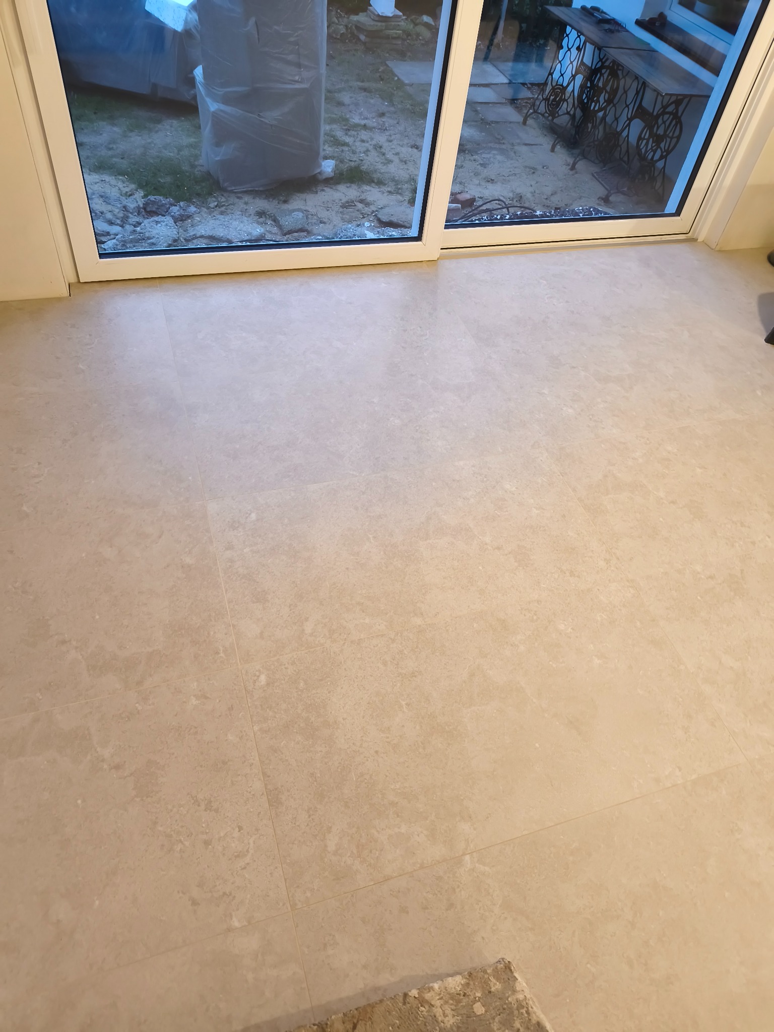 Beżowa, kamienna posadzka wewnątrz pomieszczenia, widoczna przez otwarte drzwi balkonowe. Na zewnątrz zarys stołu i roślinności. Delikatne, naturalne światło.