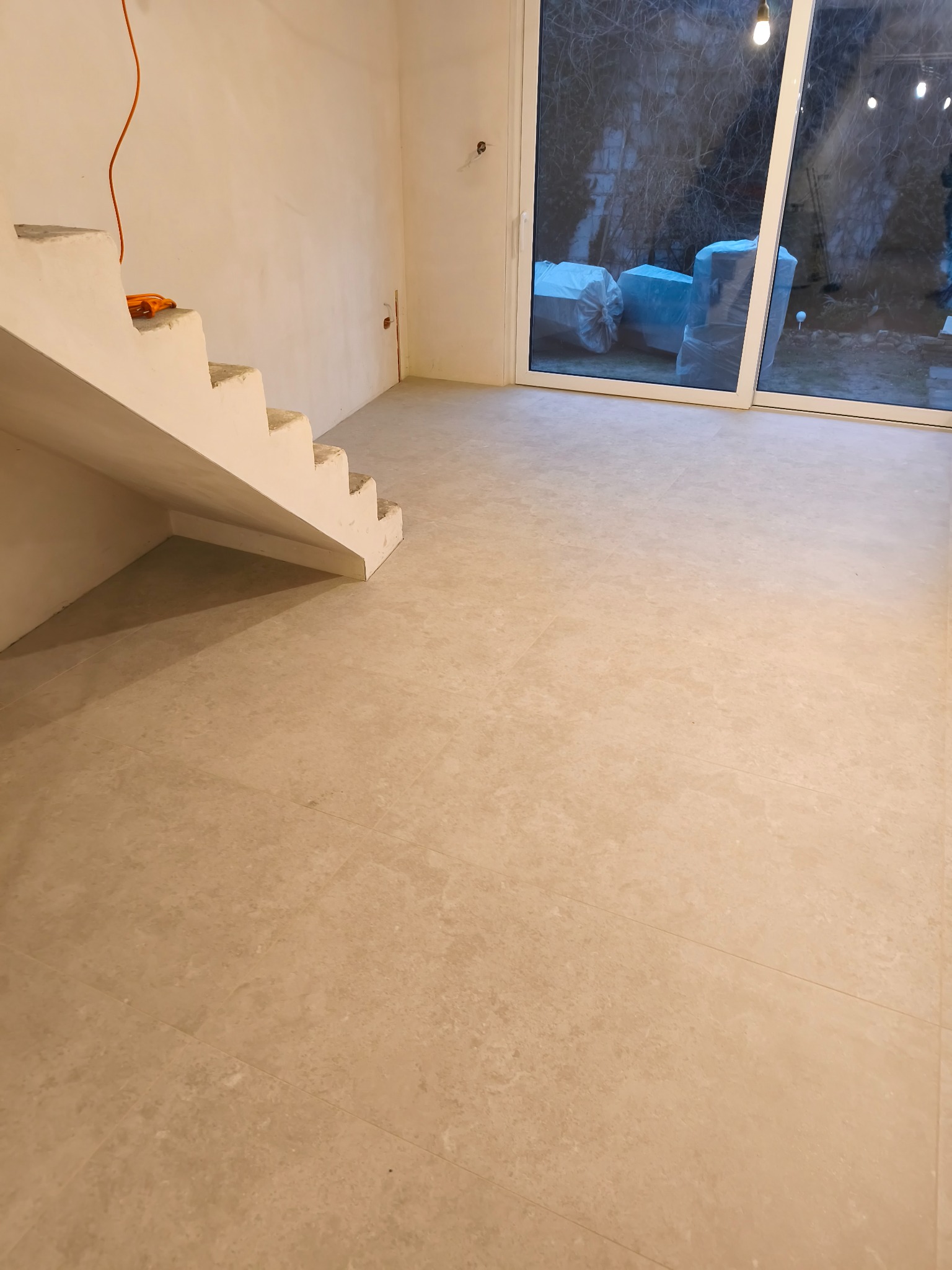 Jasne, nowo położone płytki podłogowe w przestronnym wnętrzu z betonowymi schodami i dużym oknem tarasowym. Widoczne materiały budowlane za oknem.