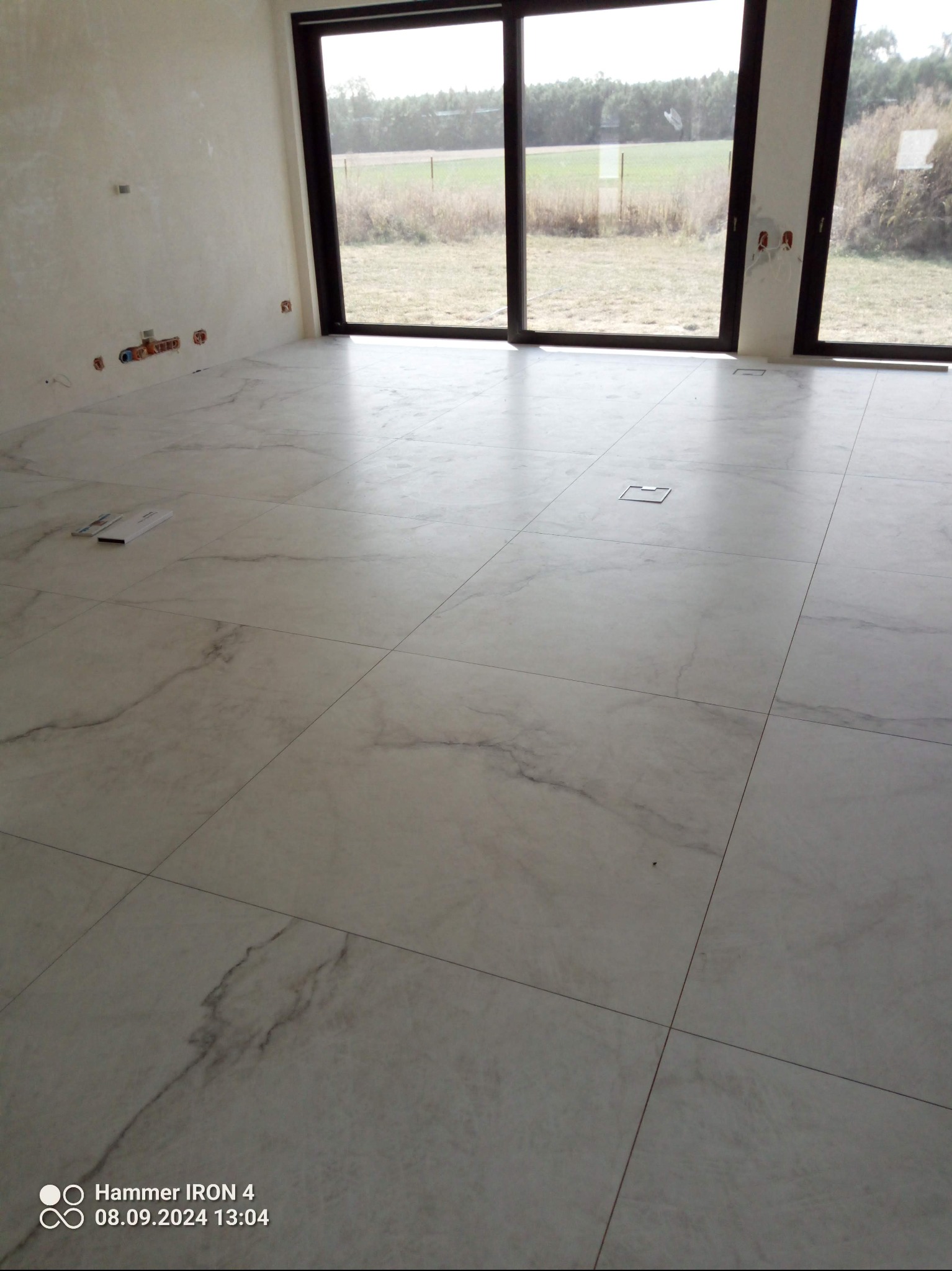 Jasne, marmurowe płytki podłogowe w nowym wnętrzu z dużym oknem. Widoczne fragmenty instalacji na ścianie i narzędzia glazurnicze. Minimalistyczny design.