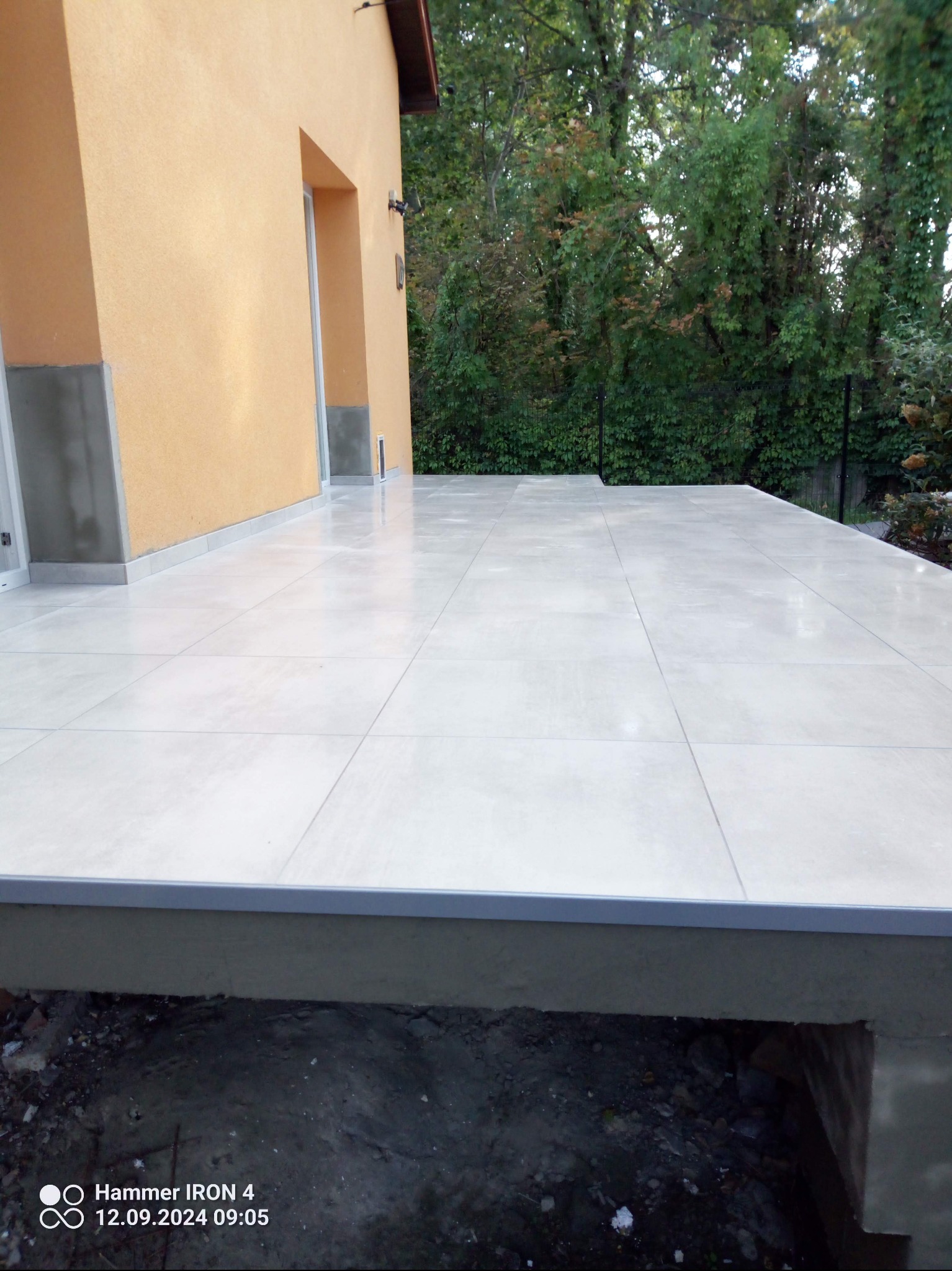 Jasne, kwadratowe płytki tarasowe ułożone na zewnątrz budynku w Warszawie. Widoczne zielone drzewa w tle. Minimalistyczny styl.