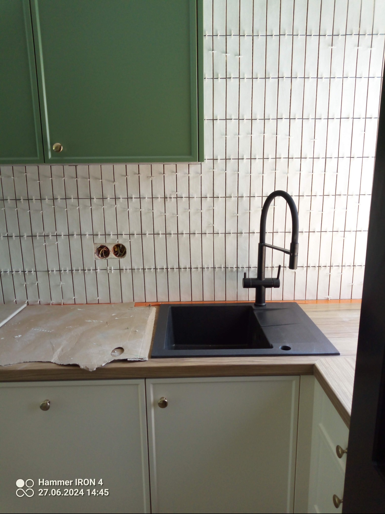 Kuchnia w trakcie wykończenia: białe, wąskie płytki ułożone pionowo na ścianie nad blatem z czarnym zlewem i zielonymi szafkami. Widoczne gniazdka elektryczne.