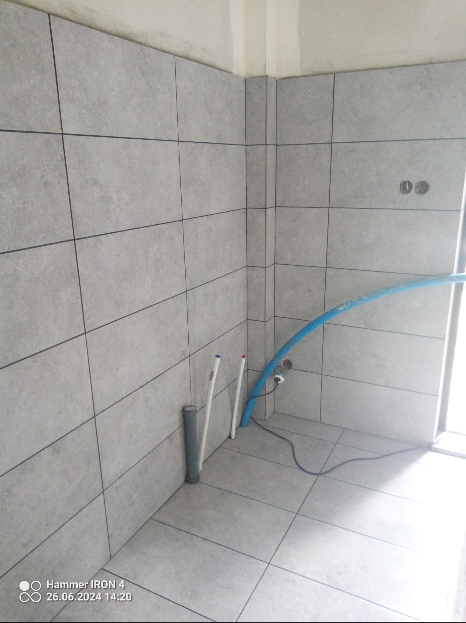 Wykończona łazienka z szarymi, matowymi płytkami na ścianach i podłodze. Widoczne rury instalacyjne i gniazdka elektryczne. Minimalistyczny design.