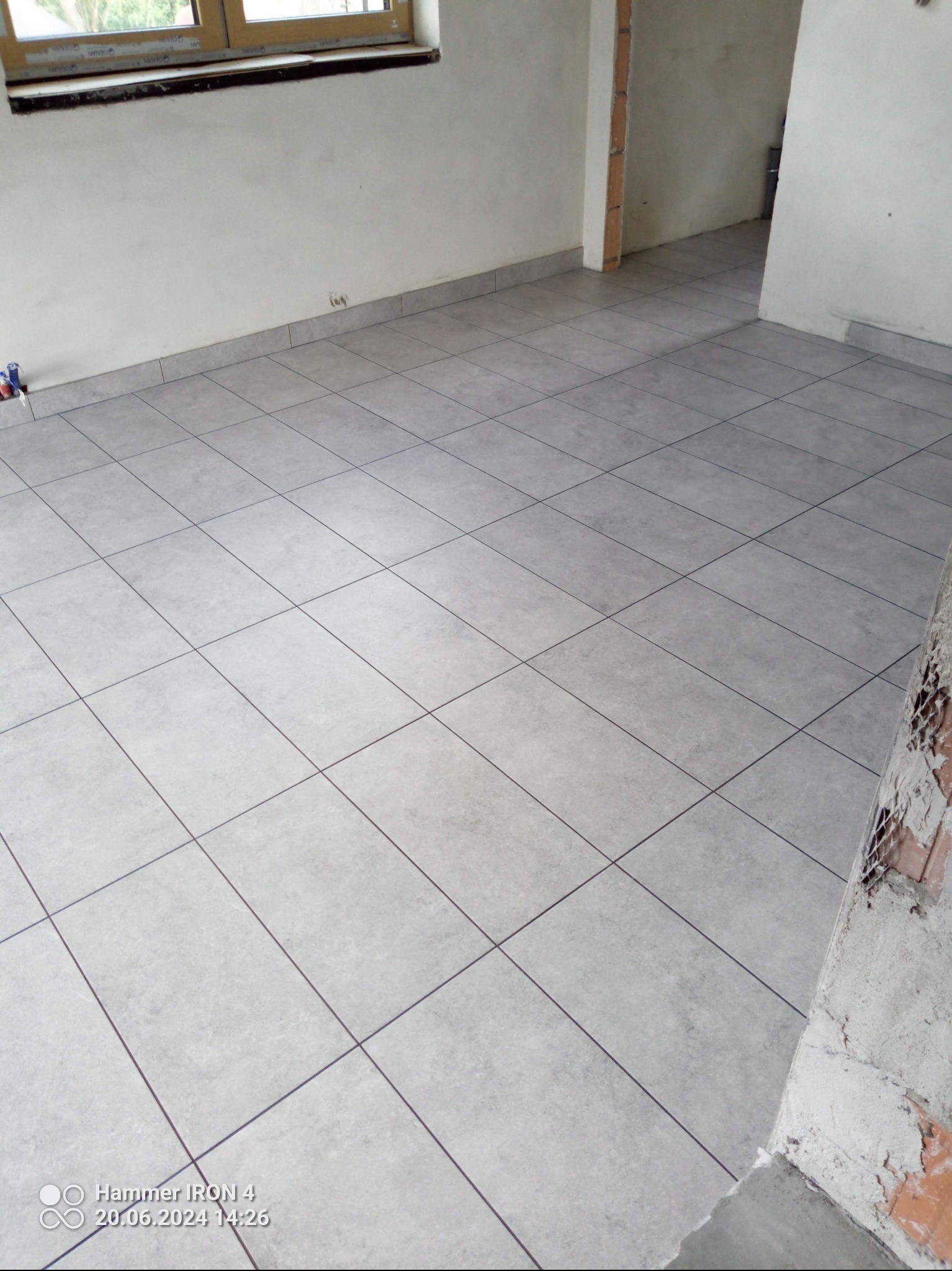 Jasnoszara, kwadratowa podłoga z płytek ceramicznych w trakcie układania, widoczne niedokończone ściany i okno. Minimalistyczny design, świeżo położona podłoga.